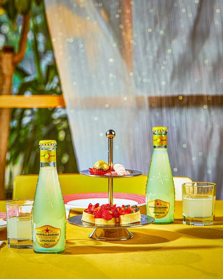 Sarı masa örtüsü üzerinde iki şişe limonata, iki katlı tatlı tepsisi ve bardaklardan oluşan masa düzeni.

