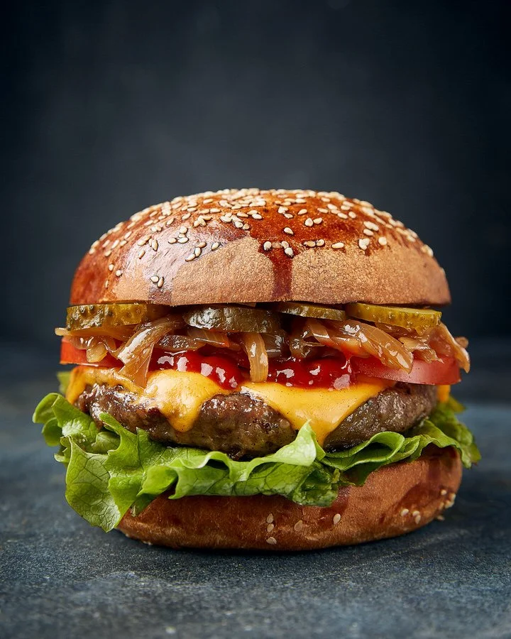 A gourmet cheeseburger with lettuce, cheese, tomatoes, caramelized onions, pickles, and a sesame seed bun.