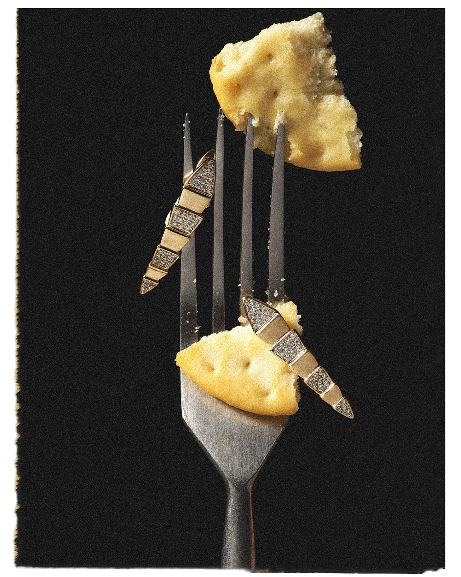 Üzerinde bir parça peynir bulunan bir çatal; altın ve elmas takılarla süslenmiş şekilde siyah bir arka plan önünde duruyor.