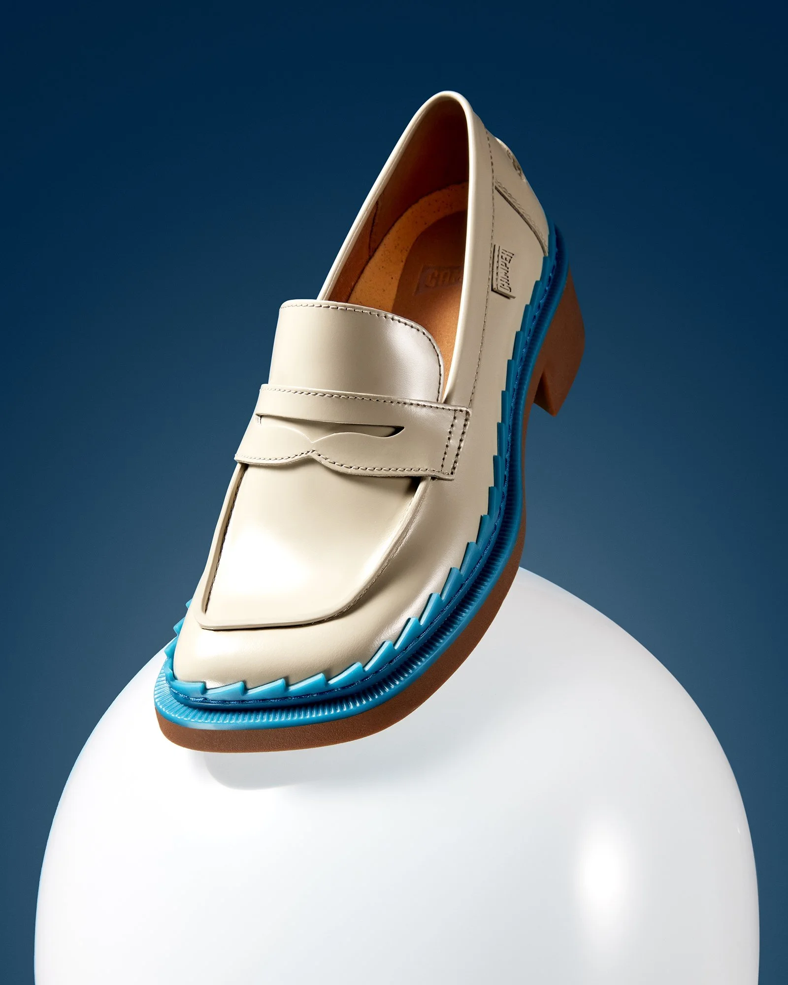 Mavi tabanlı bej deri bir loafer, koyu lacivert bir arka plan önünde beyaz yuvarlak bir kaide üzerinde sergileniyor.