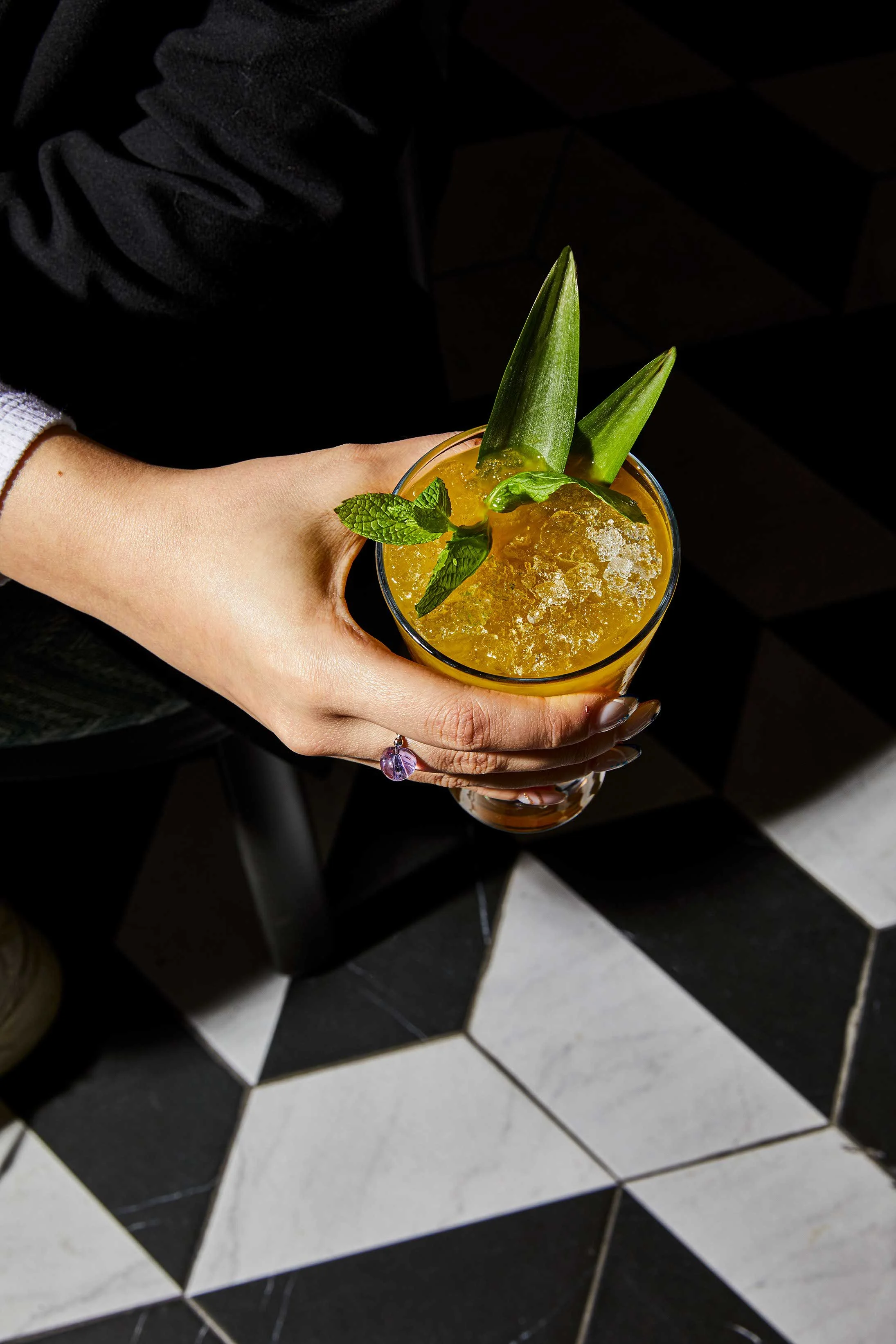 A hand holding a tropical cocktail garnished with pineapple leaves, mint, and a slice of pineapple, with crushed ice on top.