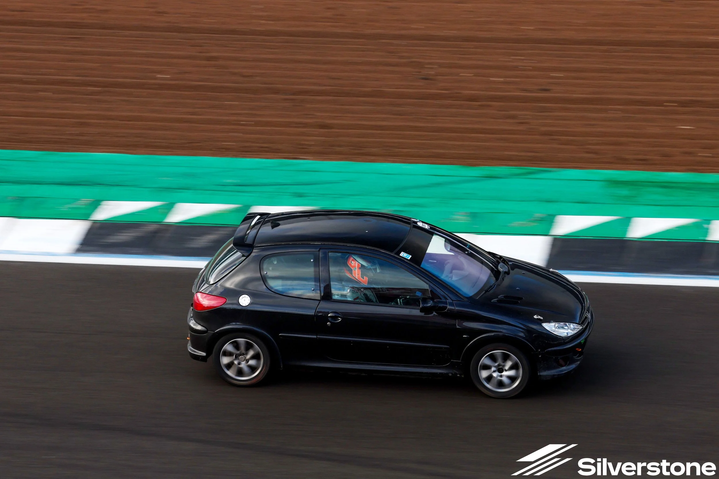 A black sports car driving on a racetrack with green and white barriers, and a brown dirt or grass area in the background.