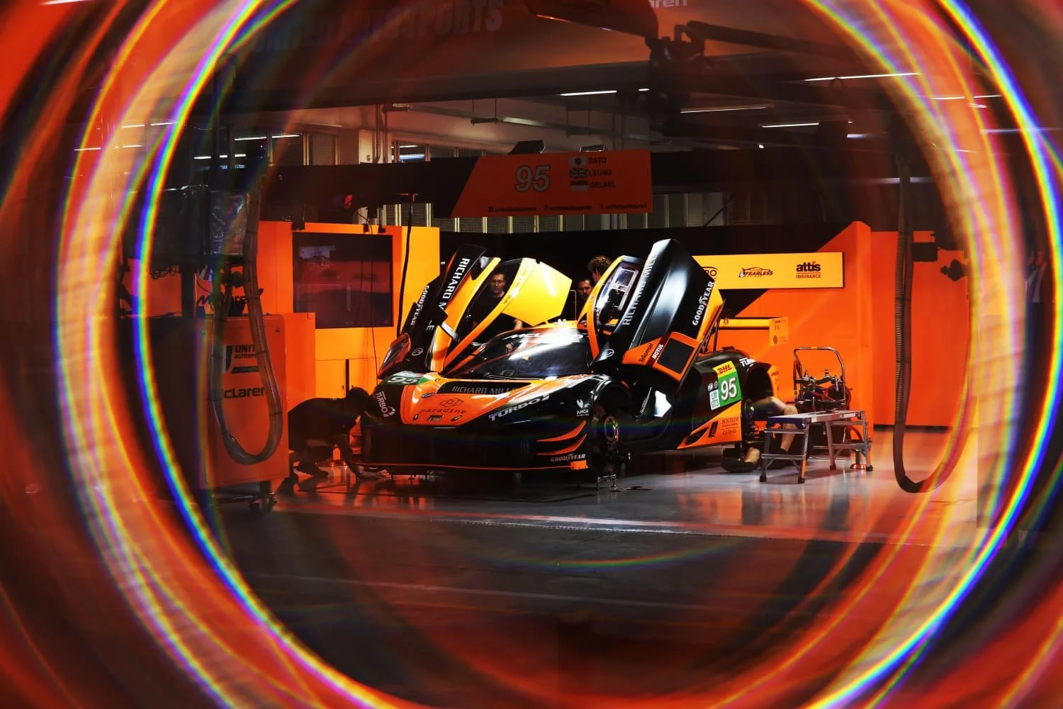 A race car in a garage with open gull-wing doors, surrounded by vibrant rainbow-colored light artifacts.