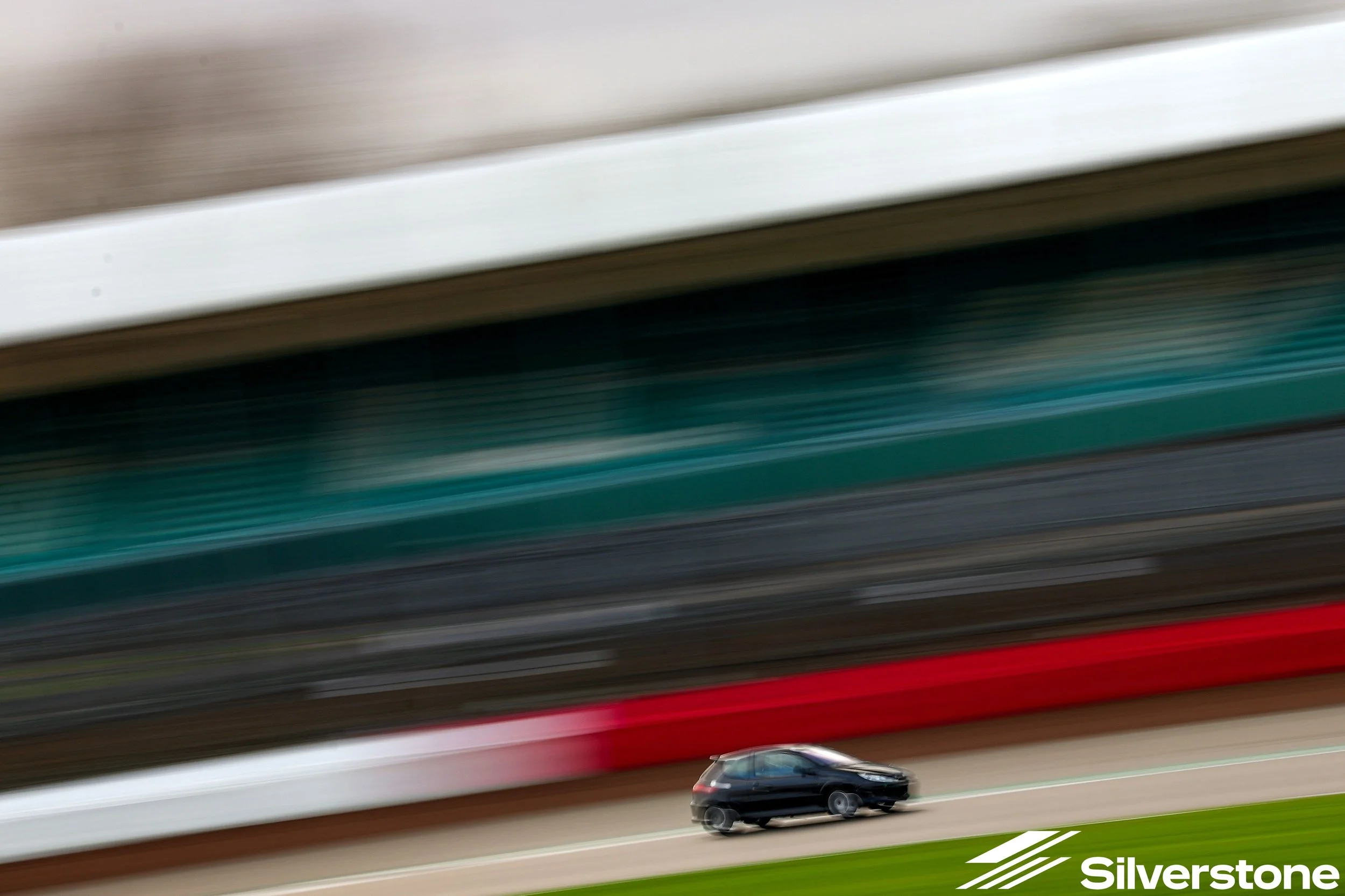 A black compact car driving on a race track with colored barriers and grandstands in the background, with motion blur suggesting high speed. Silverstone logo in the bottom right corner.