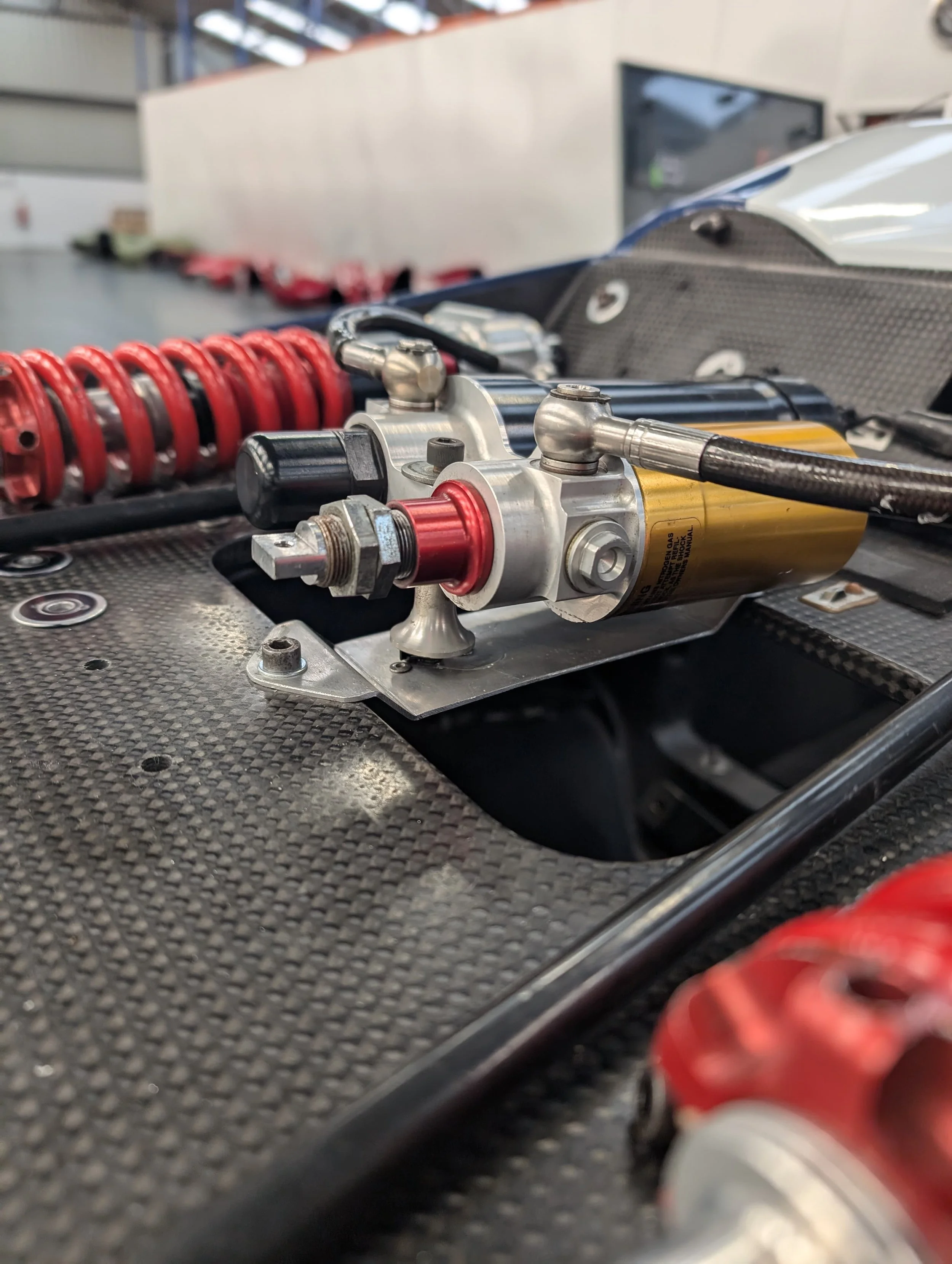 Close-up of a Formula 1 car's rear suspension and hydraulic system, with a red coil spring in the background.