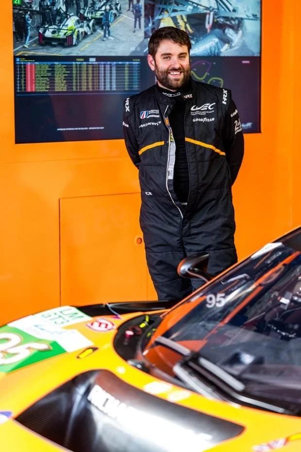 A smiling man in racing gear stands in front of a racing car with an orange backdrop and a large screen displaying a race scene in the background.