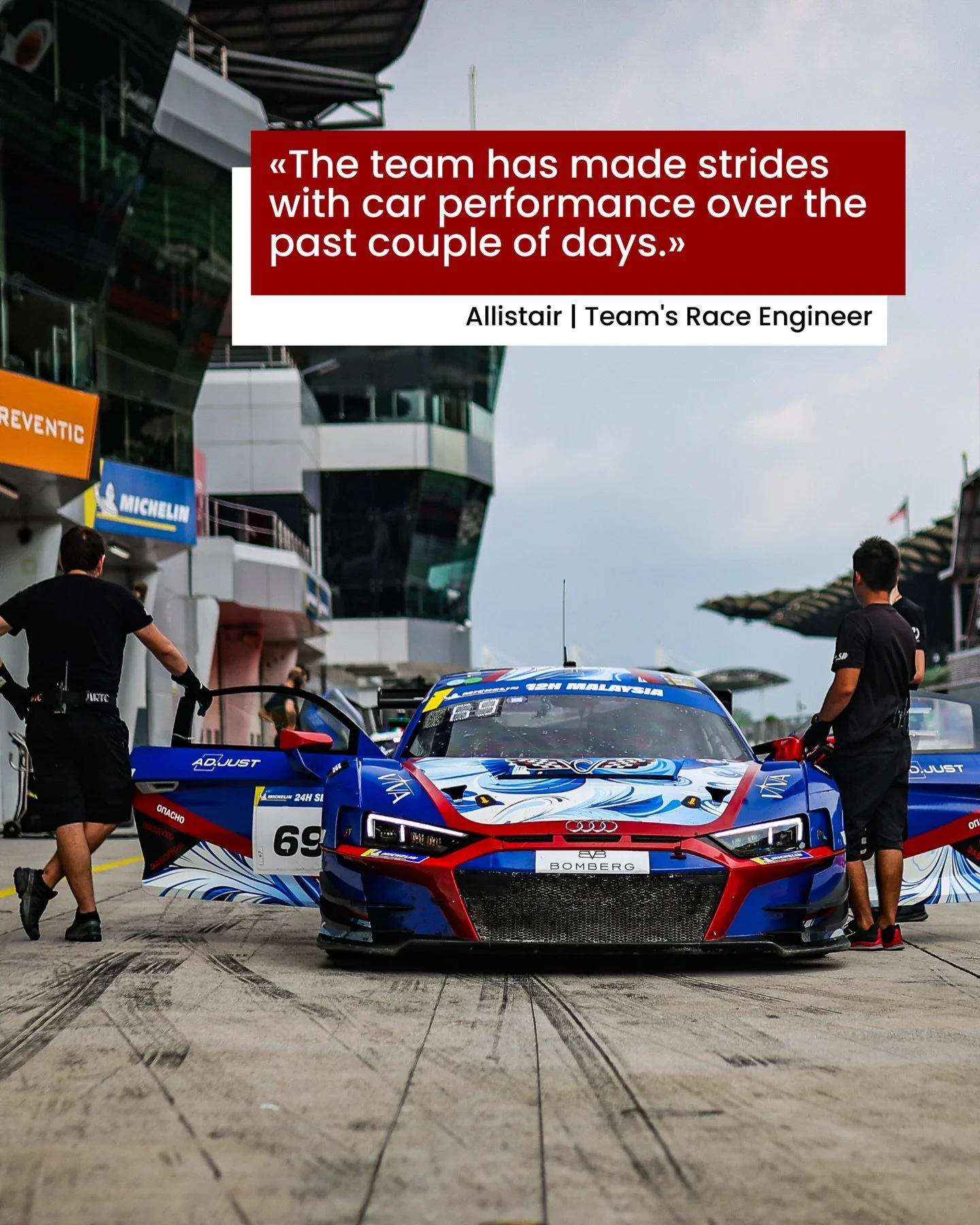 Race car in pit lane with team members around it, at a racing circuit with a modern grandstand and control tower in the background.