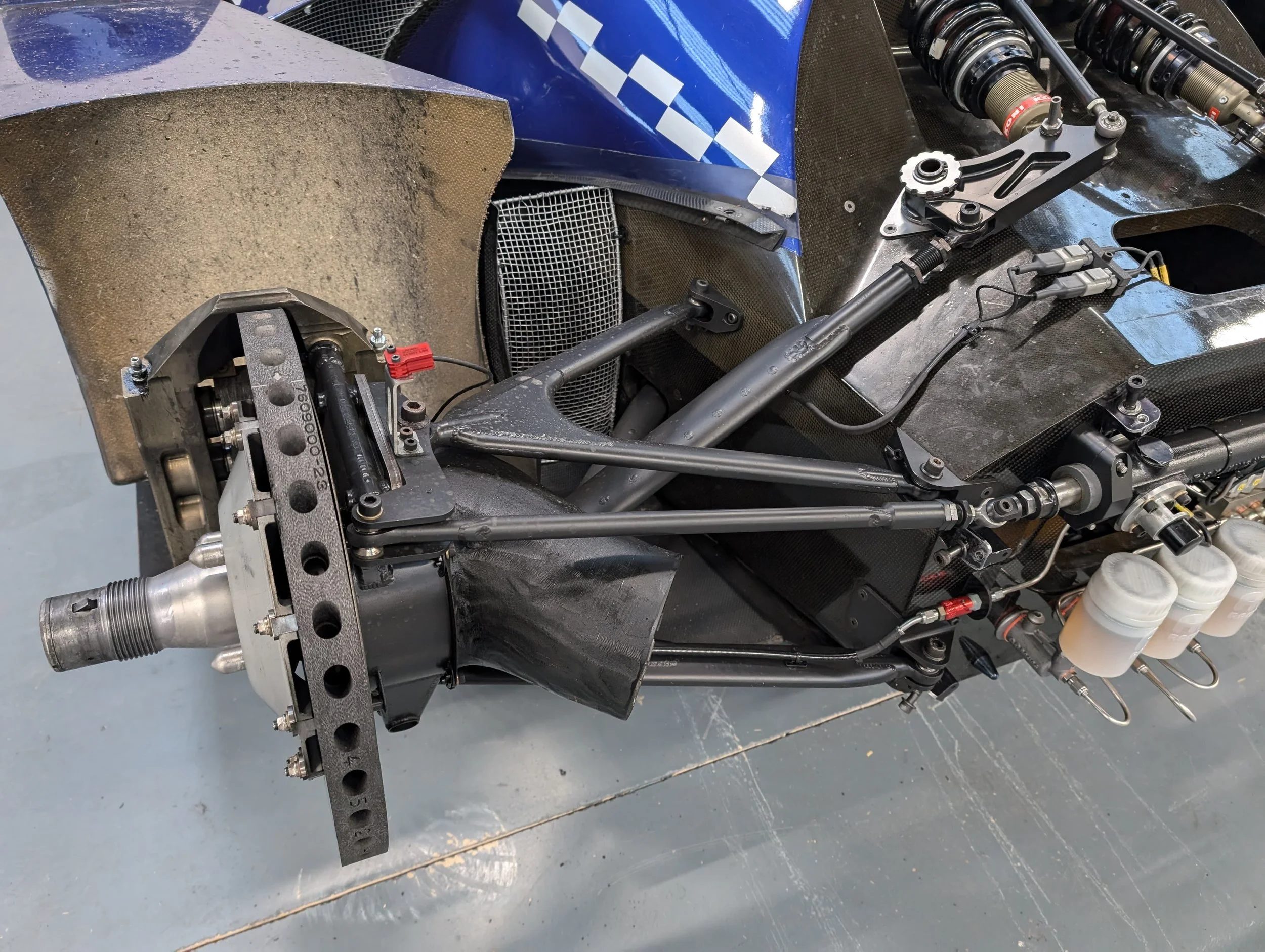 Close-up view of a high-performance race car's suspension and drivetrain components, including metal rods, shocks, and fluid reservoirs, with parts of the car's bodywork visible.