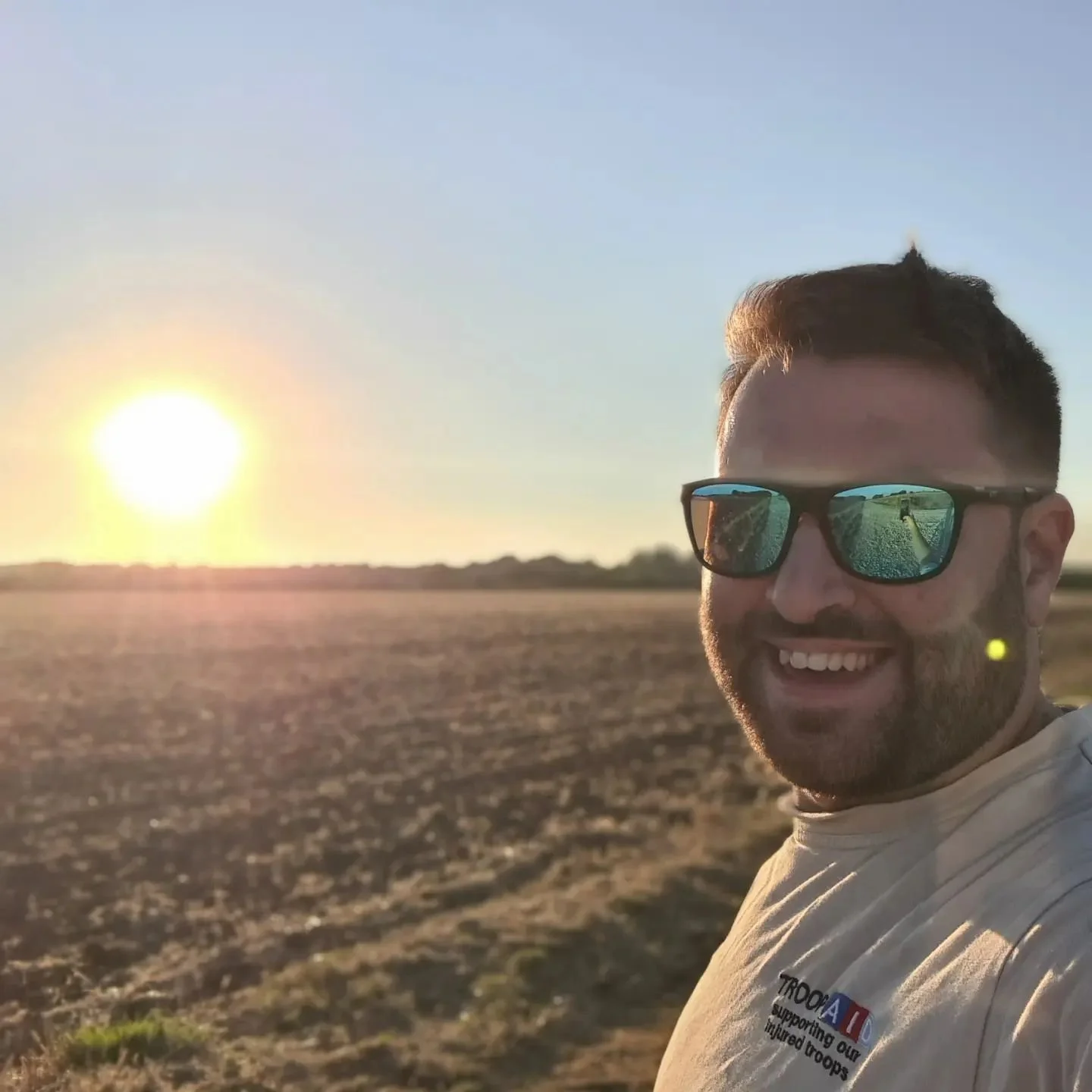 A man smiling wearing sunglasses, standing in a field at sunset with a bright sun on the horizon, wearing a white shirt.