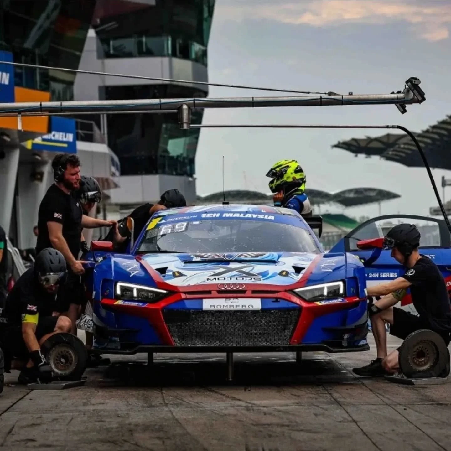 Race car in a pit stop with team members working on it, set at a race track with modern buildings and stands in the background.