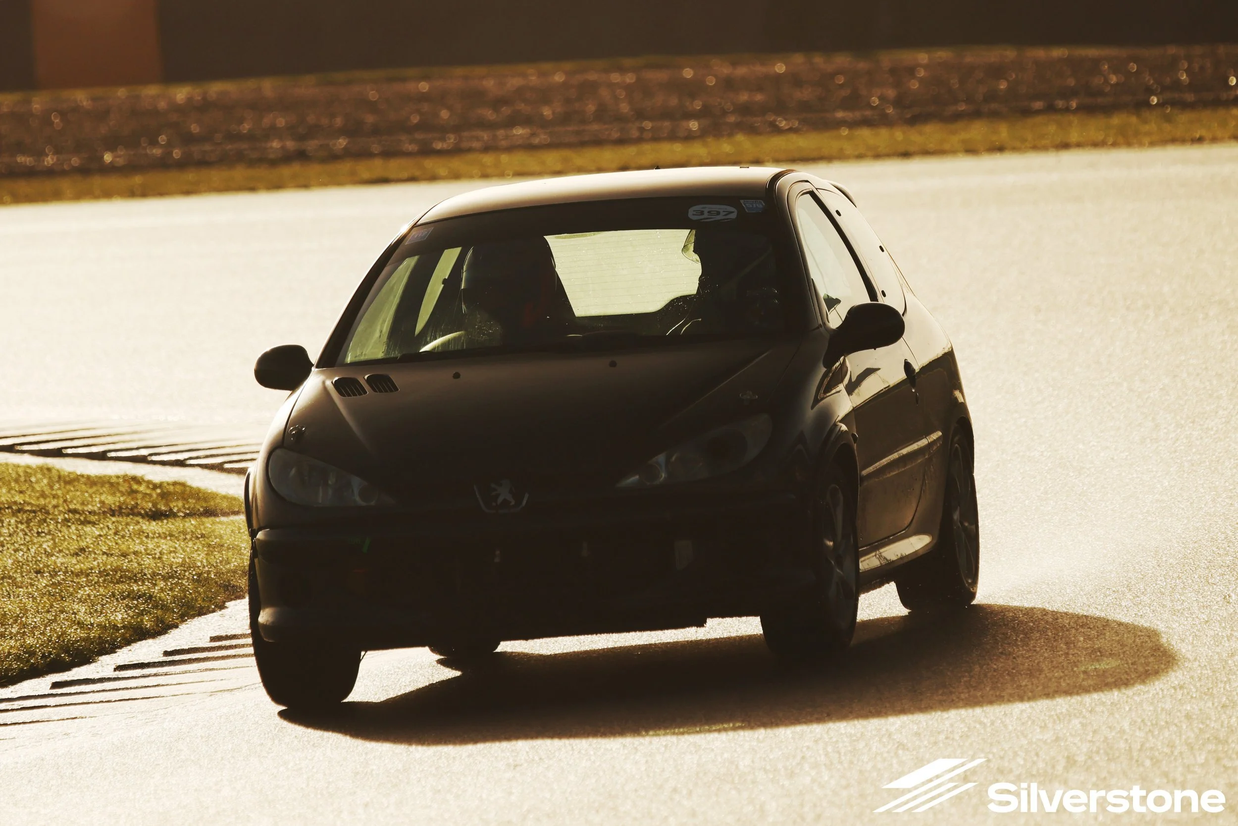 A black Peugeot compact car on a racetrack, navigating a turn with sunlight casting a shadow. The Silverstone logo is visible in the bottom right corner.