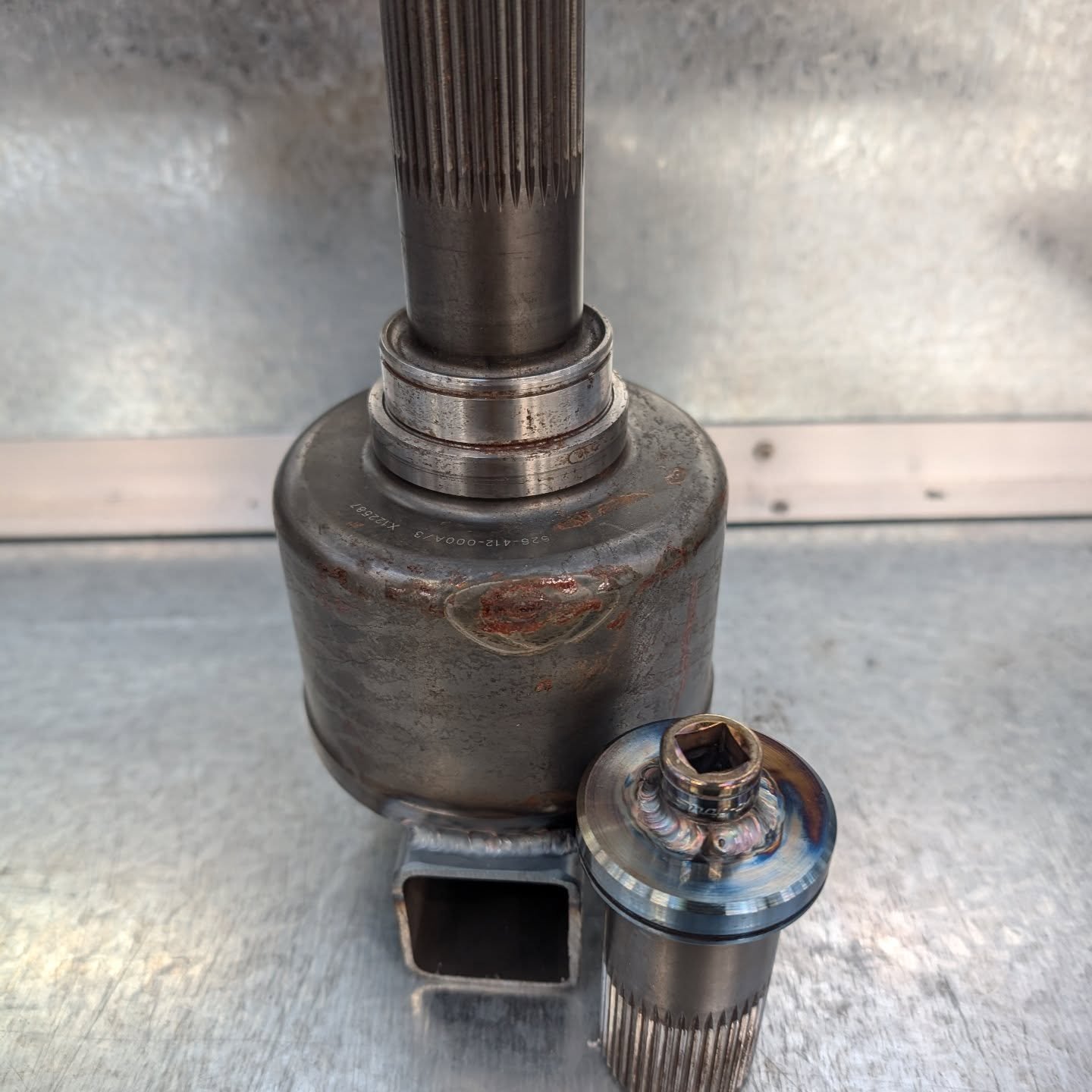 Close-up of a metal mechanical part with splined shafts and a larger cylindrical body, likely a gear or transmission component, placed on a metallic surface.