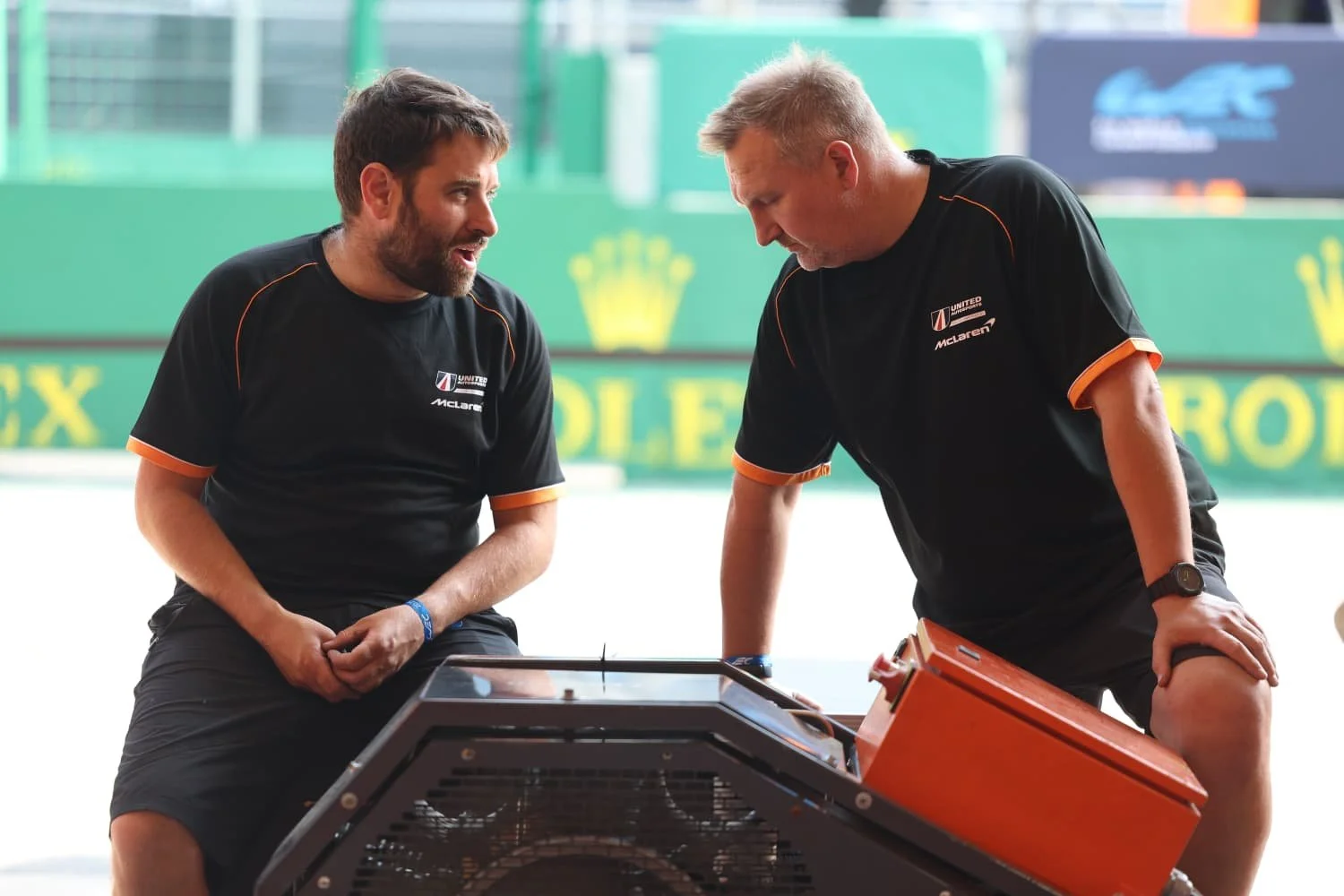 Two men in black shirts with orange trim discussing over a machine on a racetrack.