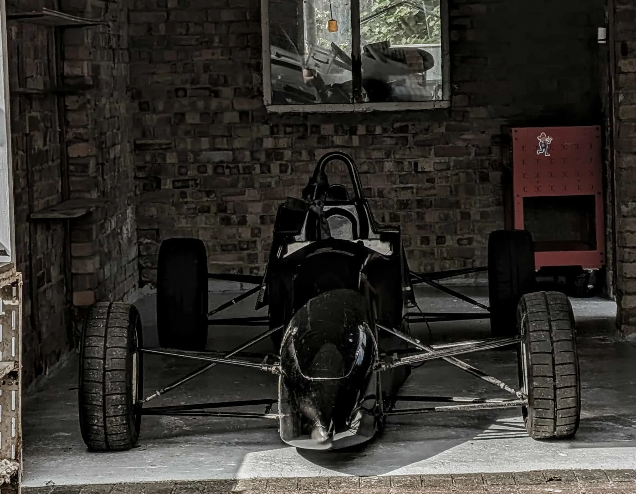 A black race car with an open cockpit and exposed wheels is parked inside a brick garage. There is a window reflecting a white vehicle outside and a red fire hydrant box on the right side.