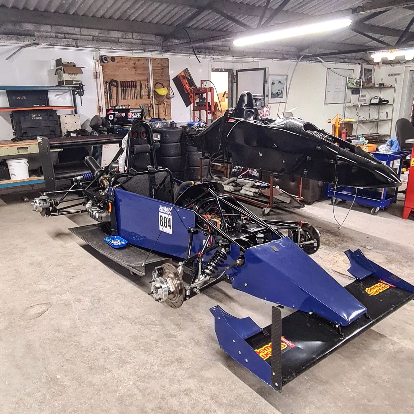 A partially assembled race car with a blue body and exposed components is inside a garage workshop. The garage has tools, tires, and equipment on the walls and shelves, with another black race car body in the background.