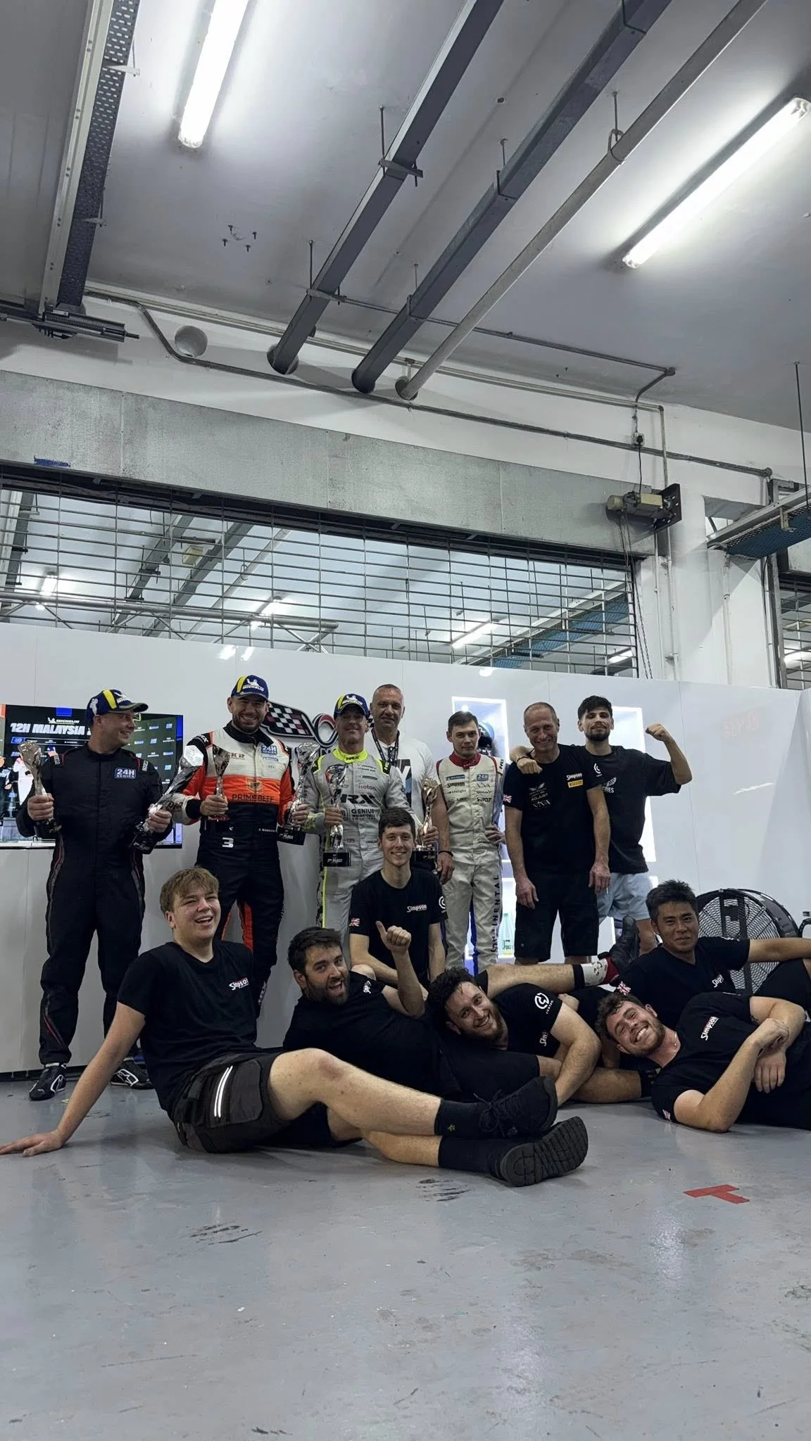 Group of race car drivers and team members celebrating after a race, some standing and some lying on the ground, in a garage or pit area.