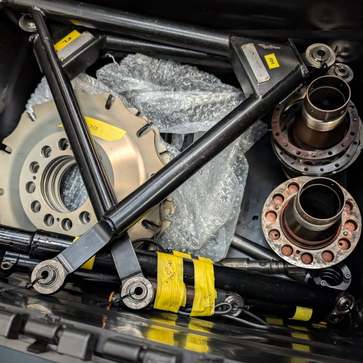 Various mechanical parts, including bearing hubs and structural components, with some wrapped in bubble wrap and yellow labels, inside a black container.