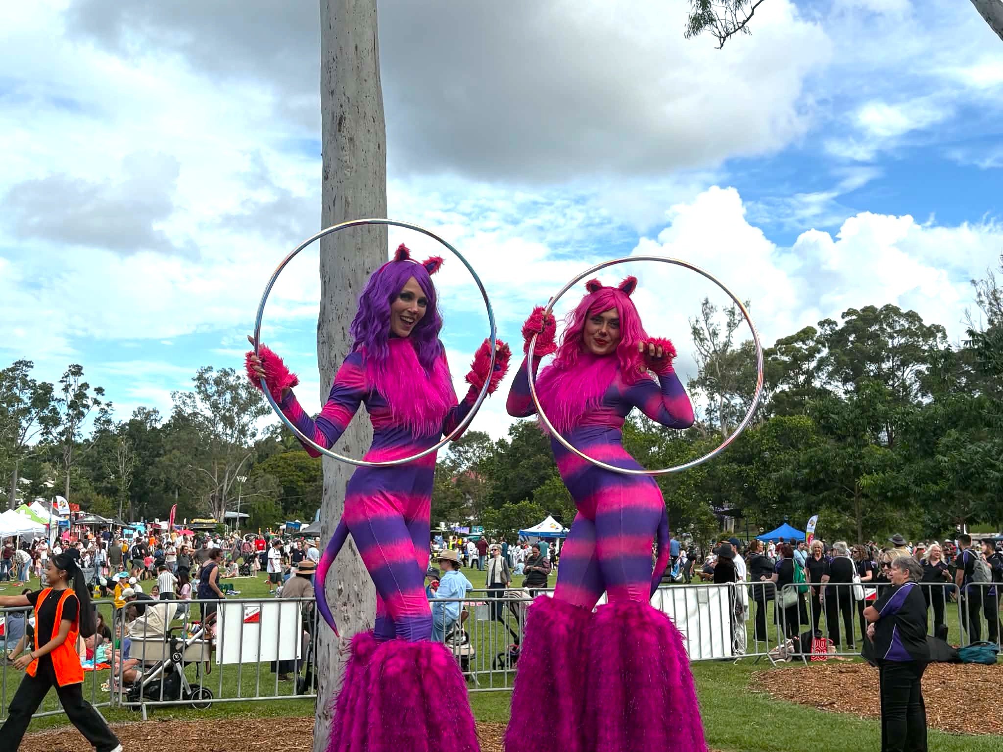 Cat-themed stilt walkers performing with hoops on grass at a busy outdoor family festival.