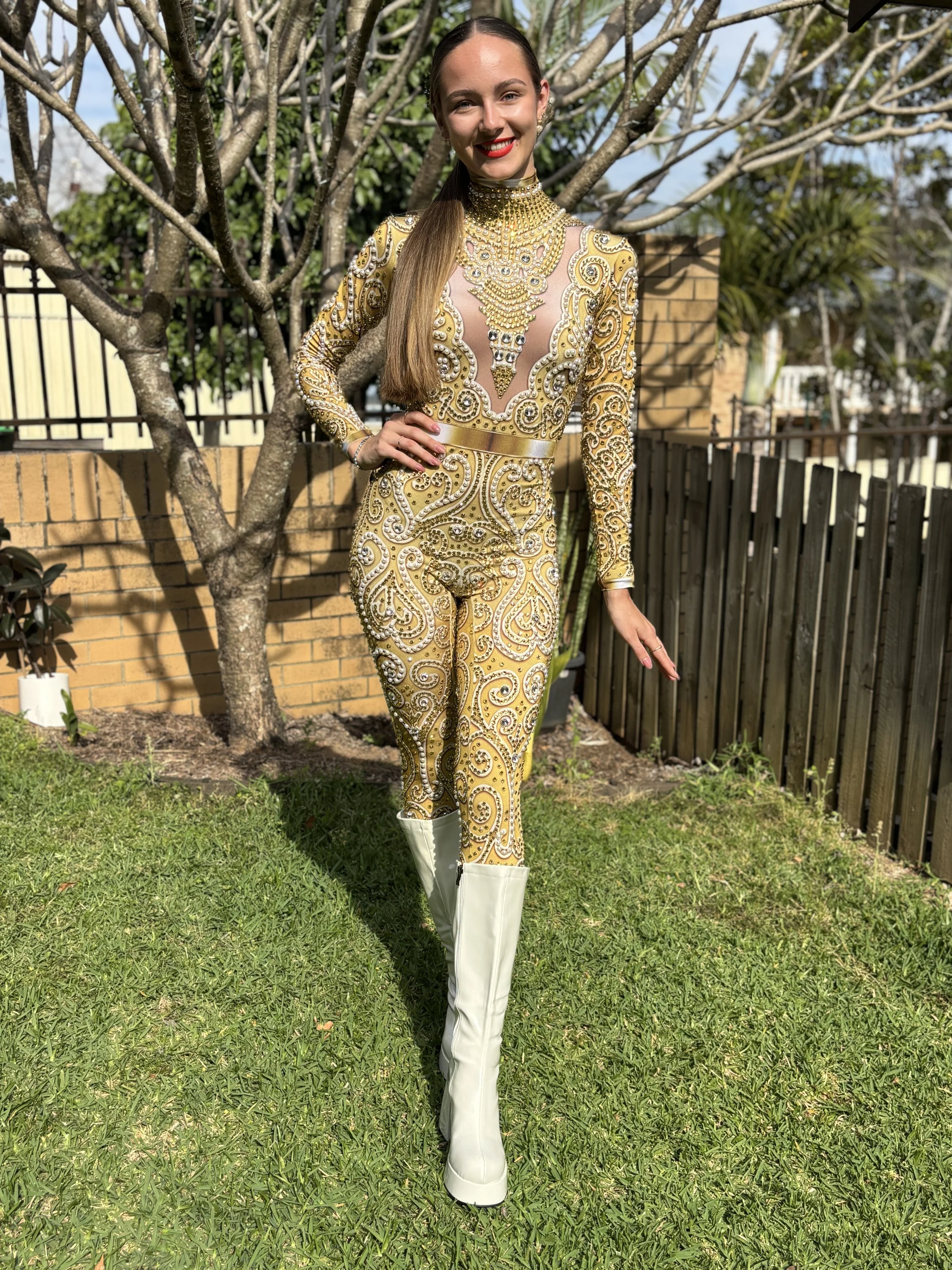 A woman in a gold event costume posing on grass with trees behind her.