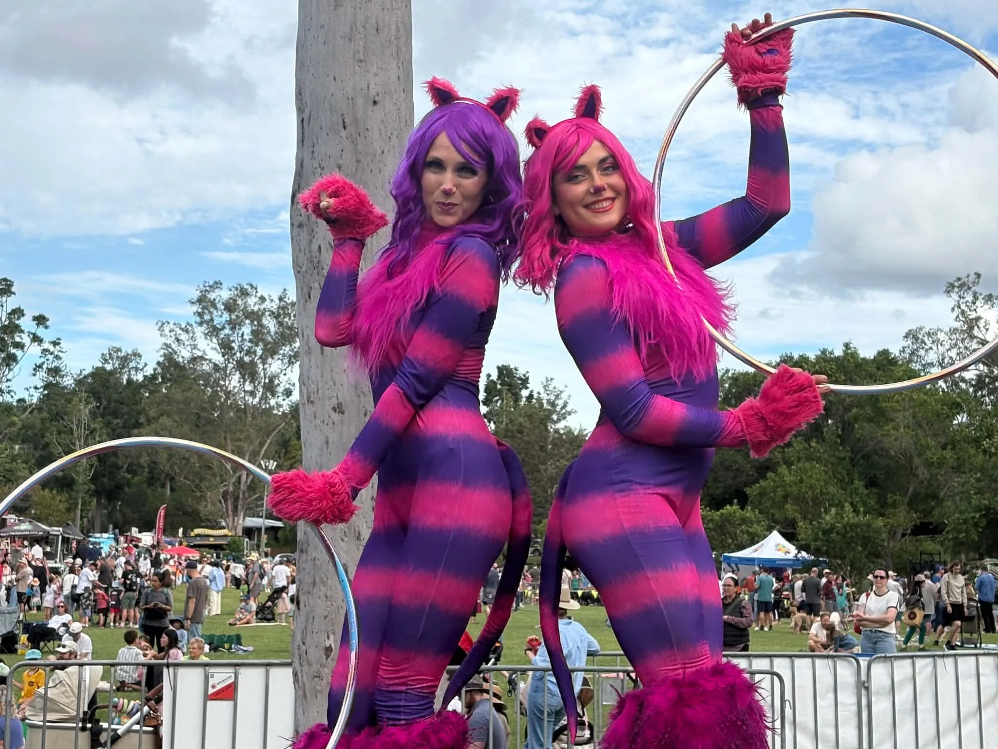 Two roving cat stilt walkers in bright pink and purple costumes entertaining crowds at a family event.