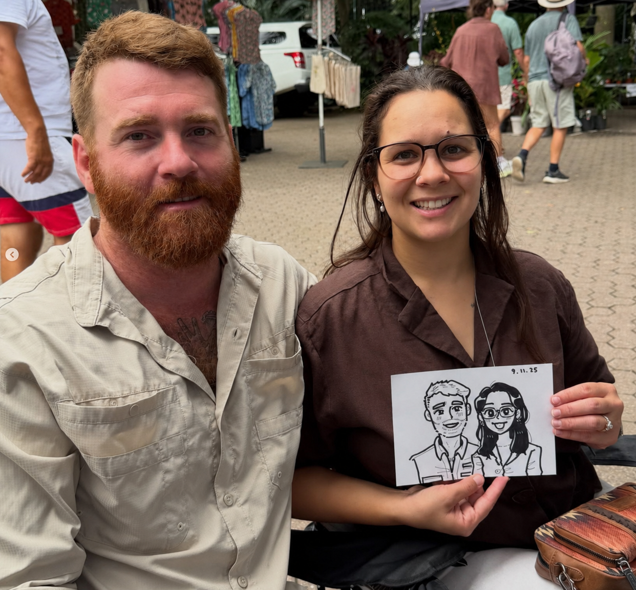 A smiling couple holding their freshly drawn 5-minute live caricature at an outdoor market event.
