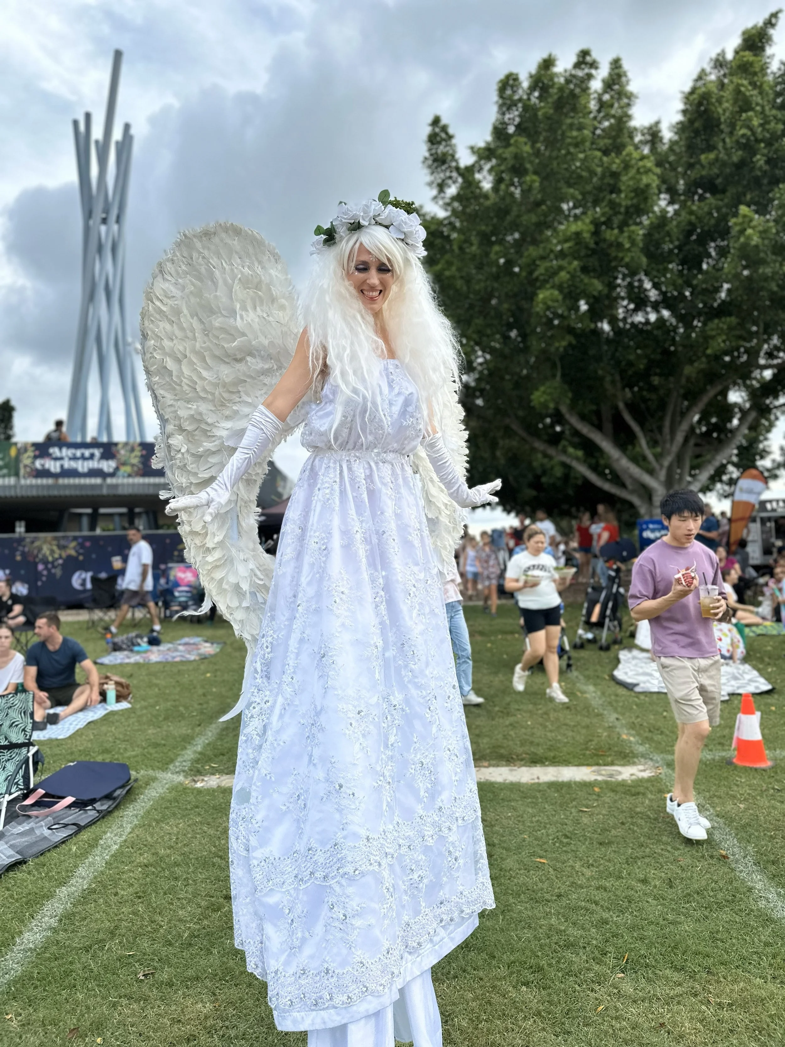 Elegant angel stilt walker with large white feather wings creating a graceful roving presence at outdoor festival.