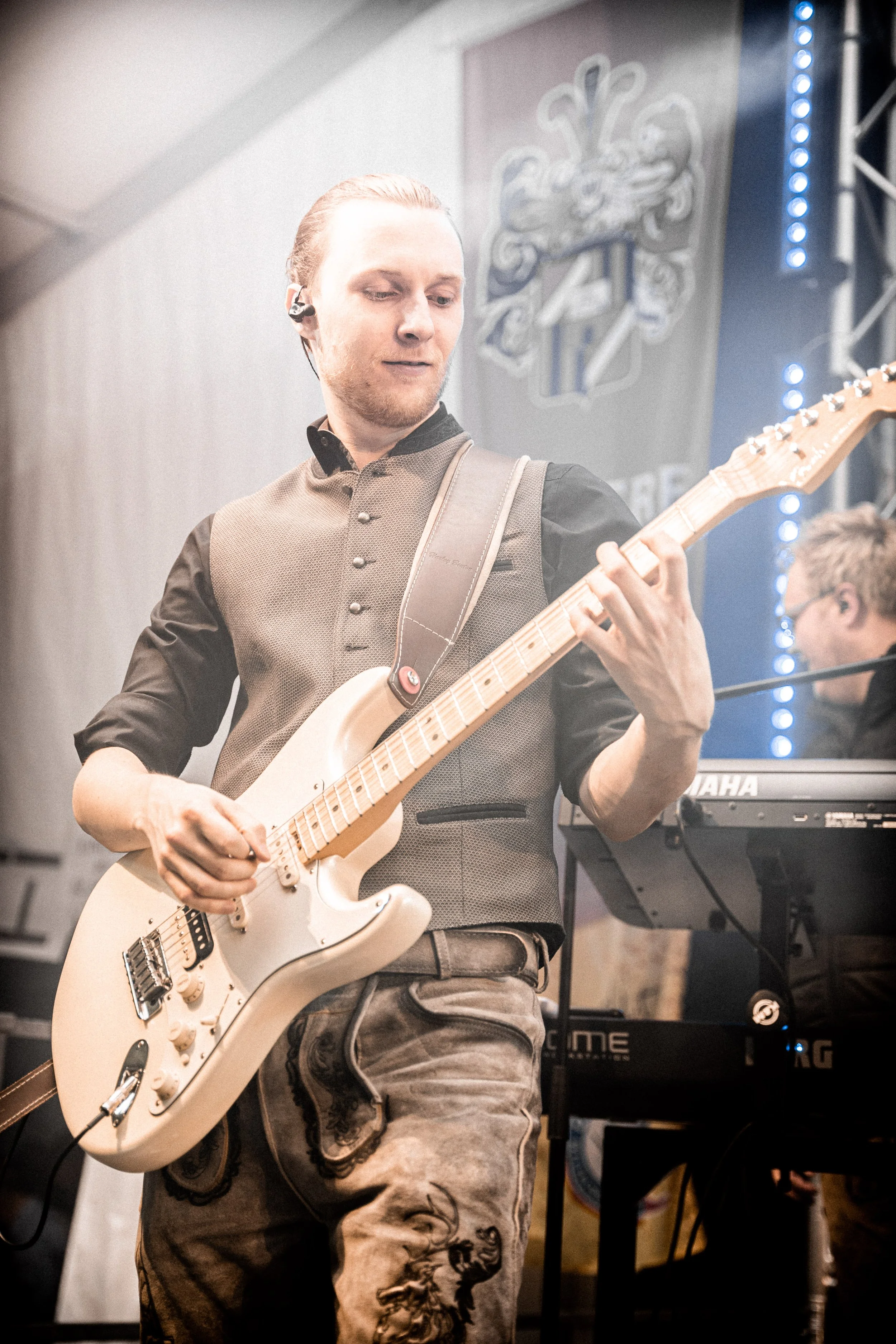 Ein Mann spielt E-Gitarre auf einer Bühne, im Hintergrund befinden sich Bühnenlichter und ein Keyboardspieler.
