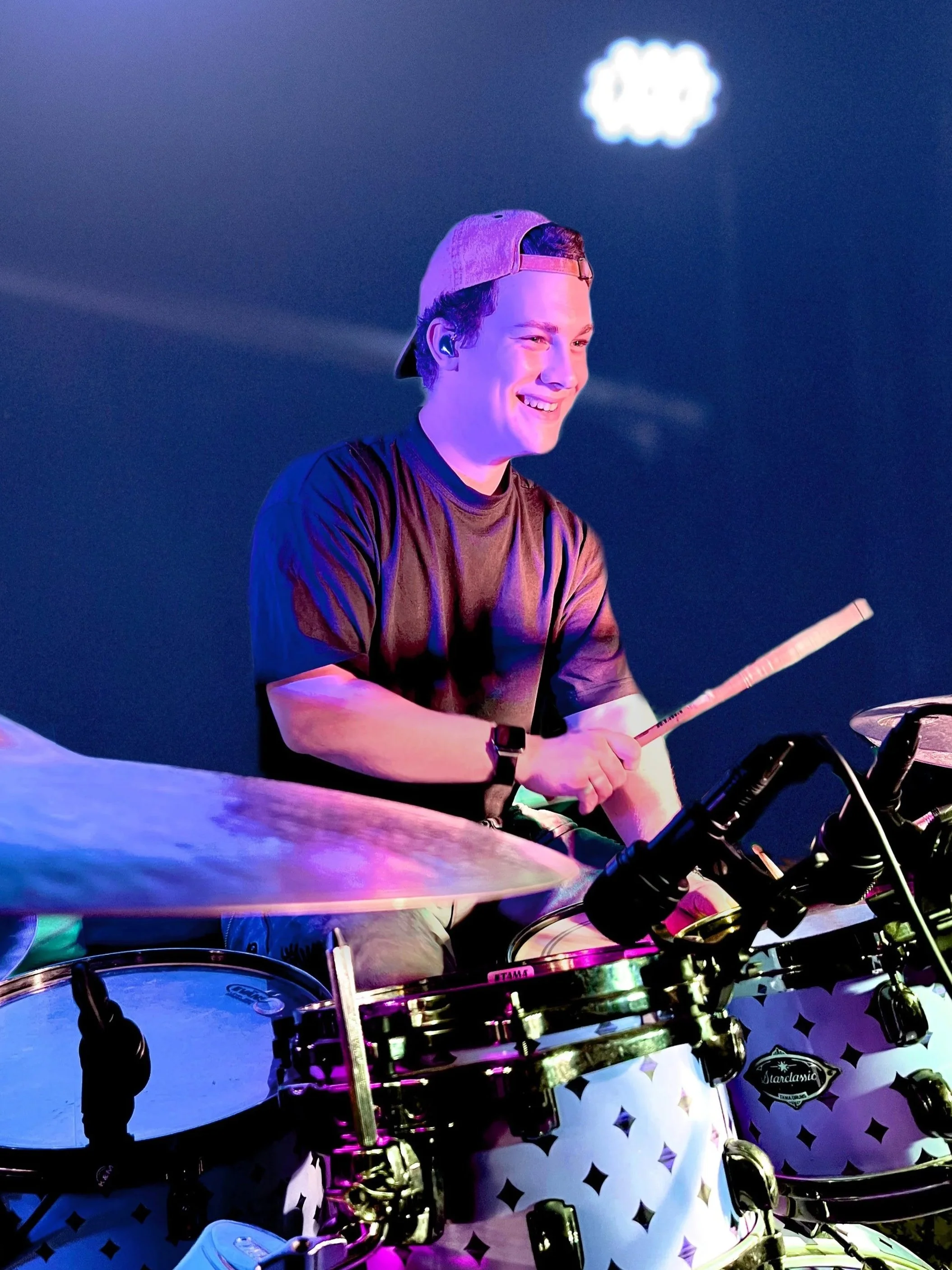 Junge Person mit Baseballkappe, lächelnd, bei einem Konzert oder Event, beleuchtet mit bunten Lichtern am Schlagzeug.