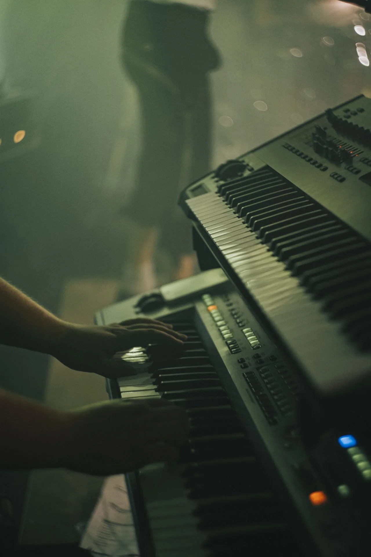 Ein Musiker spielt auf einem elektrischen Keyboard in einer dunklen, gedämpften Umgebung, während eine zweite Person im Hintergrund sichtbar ist.