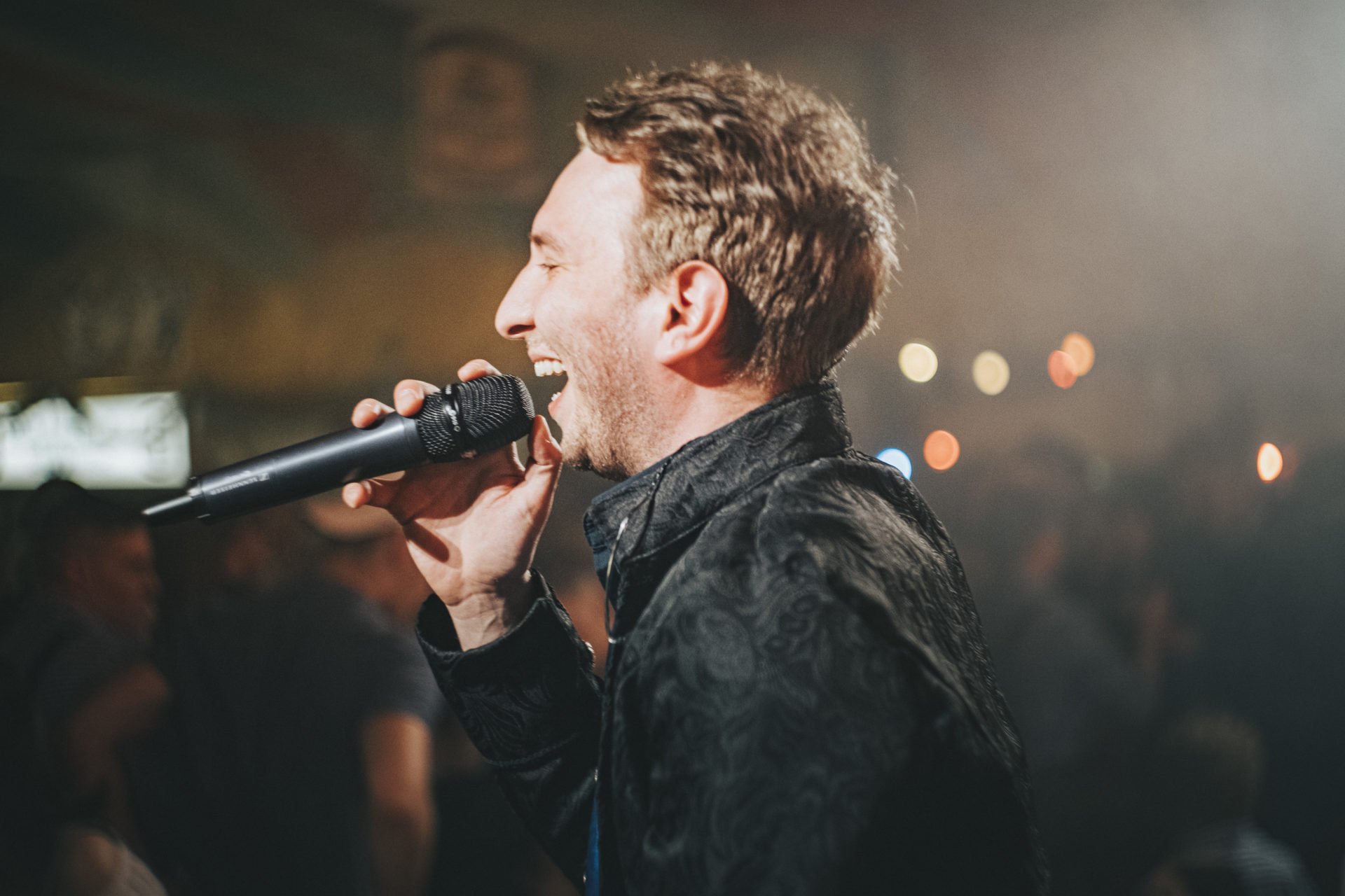 Ein lachender Mann hält ein Mikrofon in der Hand und singt auf einer Bühne, während im Hintergrund unscharfe Lichter zu sehen sind.