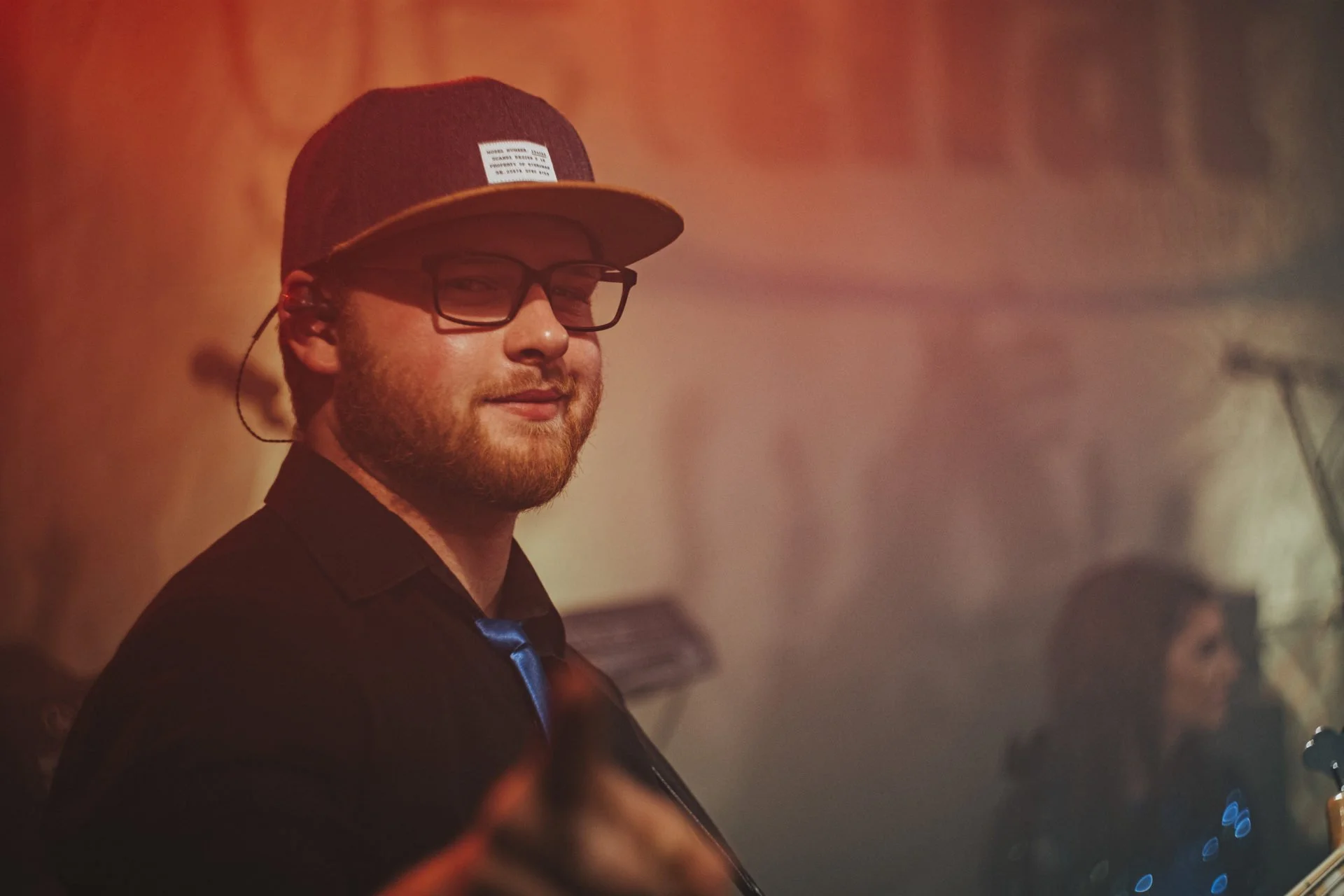 Ein junger Mann mit Brille, Hut und dunklem Hemd, der in einem Raum mit rotem Licht steht.