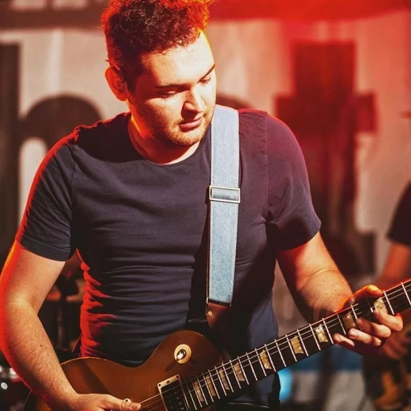 Ein junger Mann spielt Gitarre auf einer Bühne bei rotem Licht.