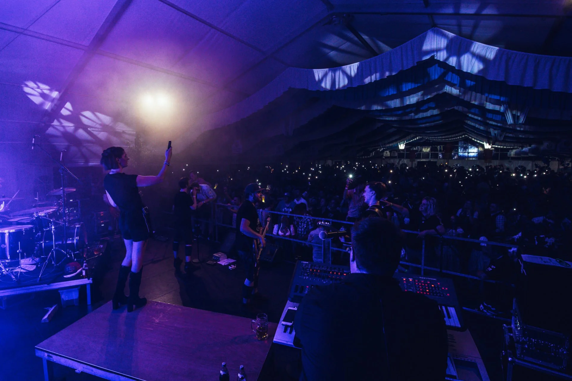 Bühne mit einer Live-Band während eines Konzerts in einem Zelt, im Vordergrund sind Musiker und ein Publikum mit vielen leuchtenden Handys