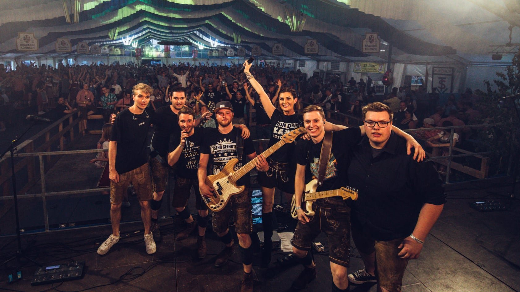 Gruppe von Musikern auf einer Bühne, mit Publikum im Hintergrund bei einem Konzert in einem Festzelt.