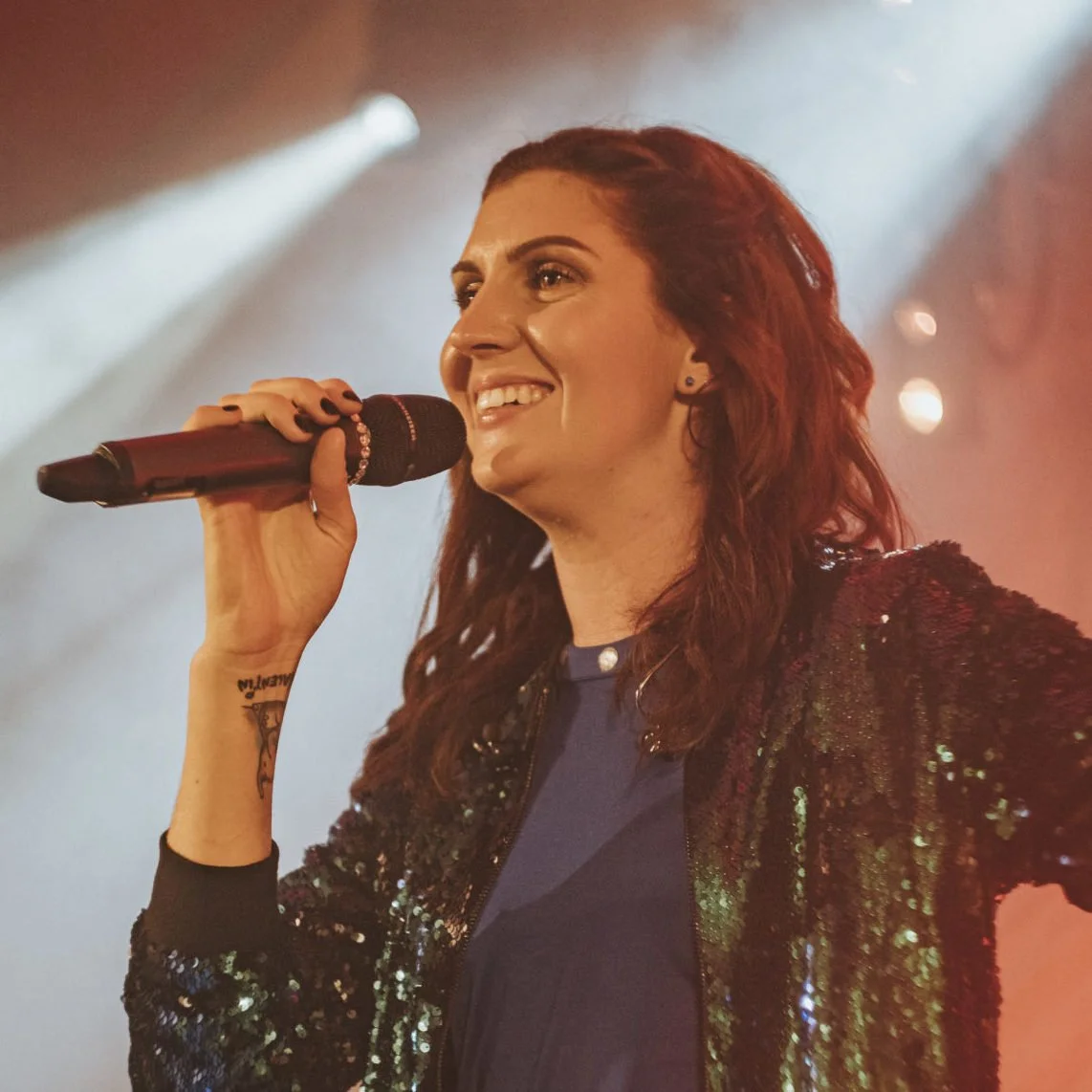 Eine junge Frau mit braunen, lockigen Haaren hält ein Mikrofon beim Singen auf einer Bühne, im Hintergrund Scheinwerferlicht und Rauch.