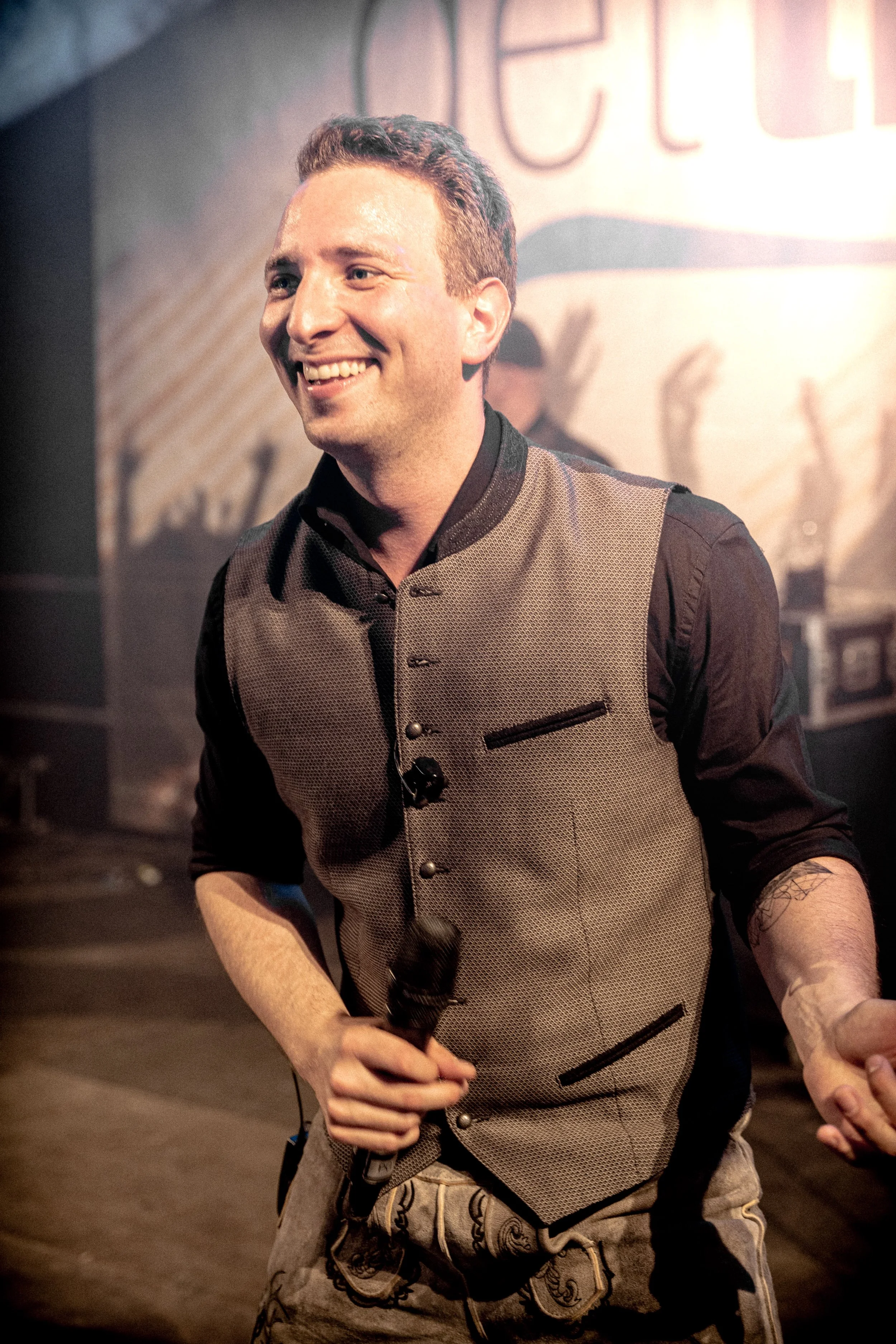 Ein lachender Mann hält ein Mikrofon in der Hand, trägt eine Weste und ist auf einer Bühne, im Hintergrund graffitiartige Kunst.