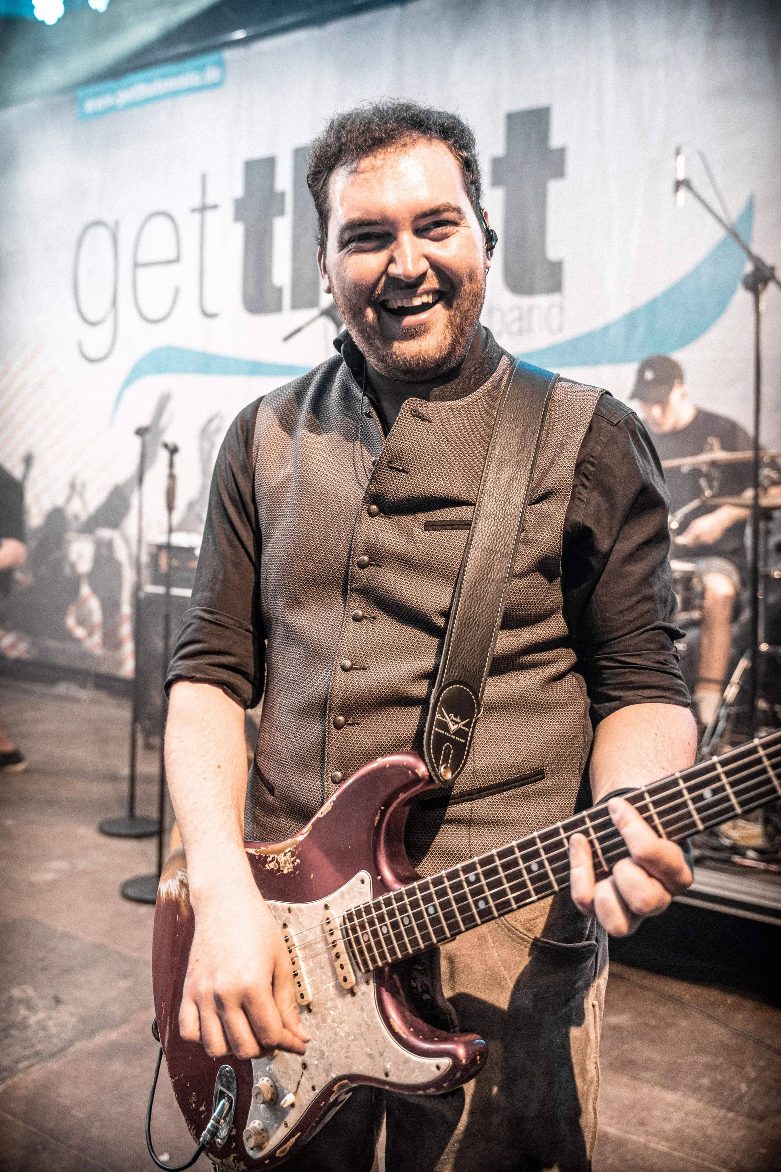 Ein lachender Musiker spielt E-Gitarre auf Bühne bei einem Konzert, im Hintergrund sind andere Bandmitglieder und ein Banner mit dem Logo 'get<heart>t'.