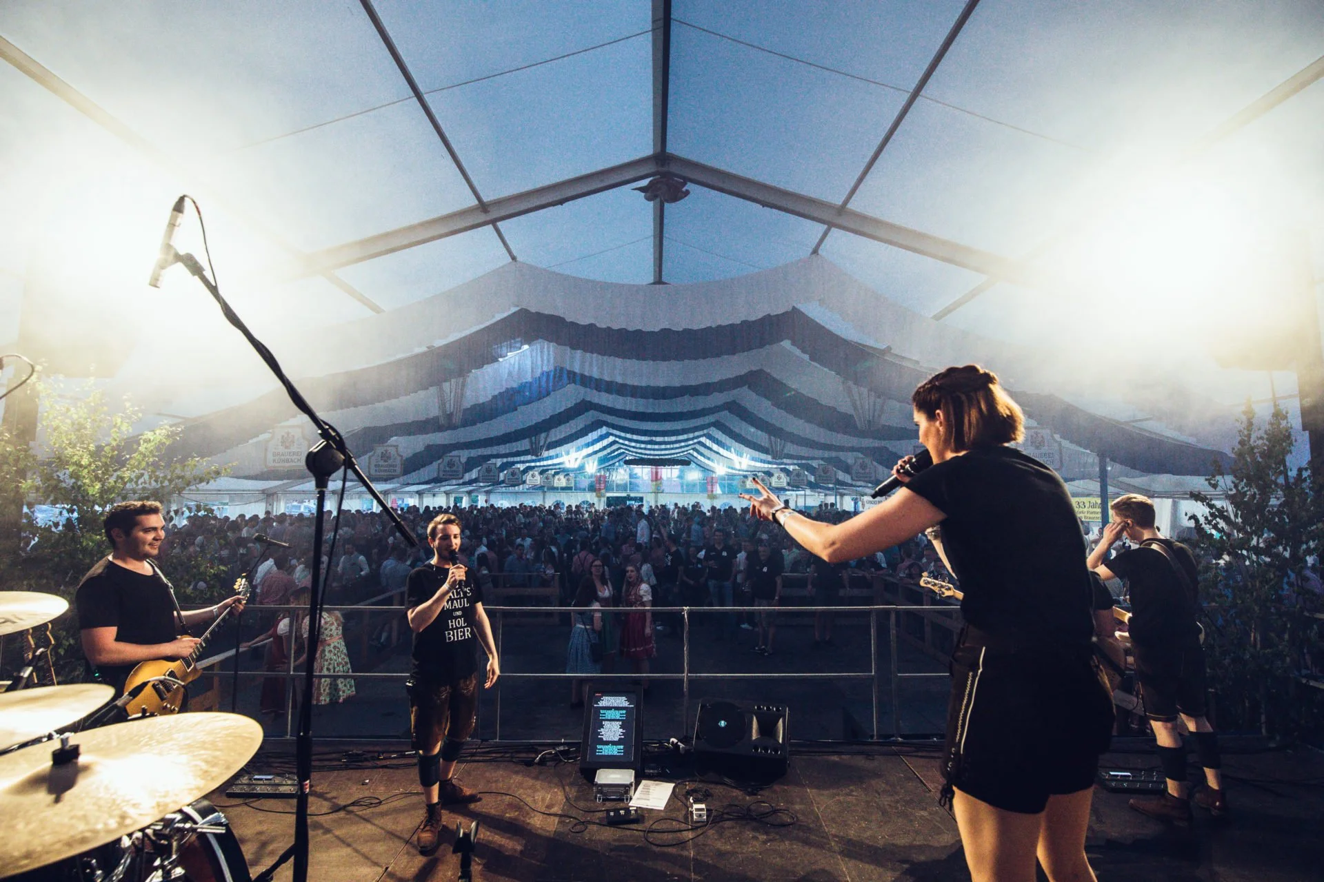 Musiker auf einer Bühne bei einem Festival im Zelt, im Hintergrund ein Publikum unter weißen und blauen Fahnen, mit einem Musikständer, Mikrofon, Gitarren und Schlagzeug.
