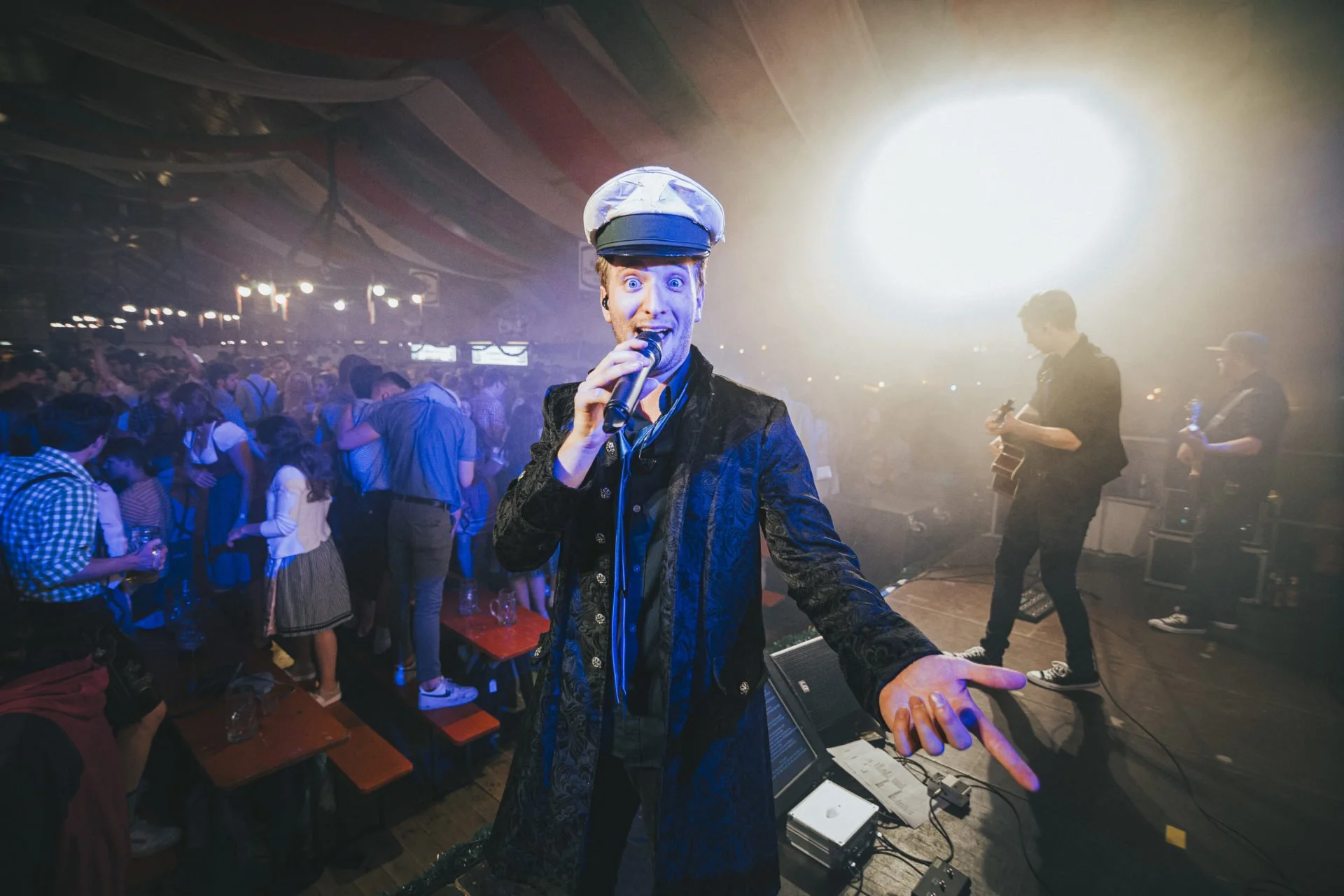 Ein Sänger in Kapitänsmütze singt in einer Band auf der Bühne, während hinten zwei Musiker Gitarre spielen, in einer belebten partyähnlichen Umgebung mit Tanzenden.