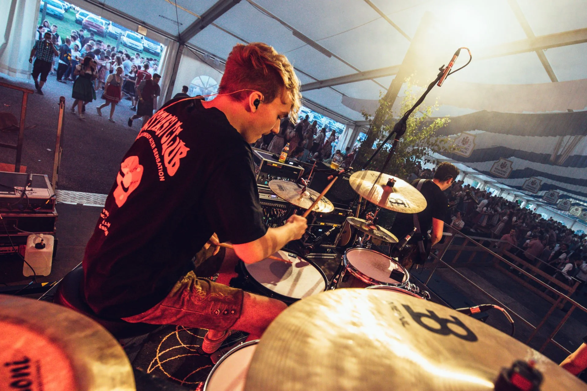 Musiker spielt Schlagzeug auf einer Bühne in einer großen Festhalle, im Hintergrund viele Menschen beim Festzelt in Bayern.