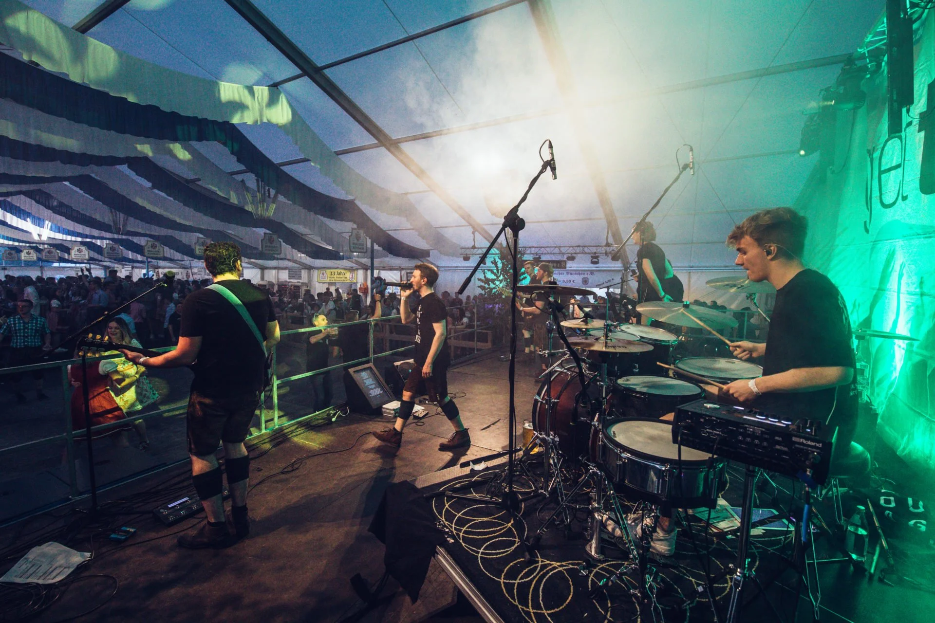 Band spielt auf einer Bühne in einem festlich geschmückten Zelt mit Publikum im Hintergrund.