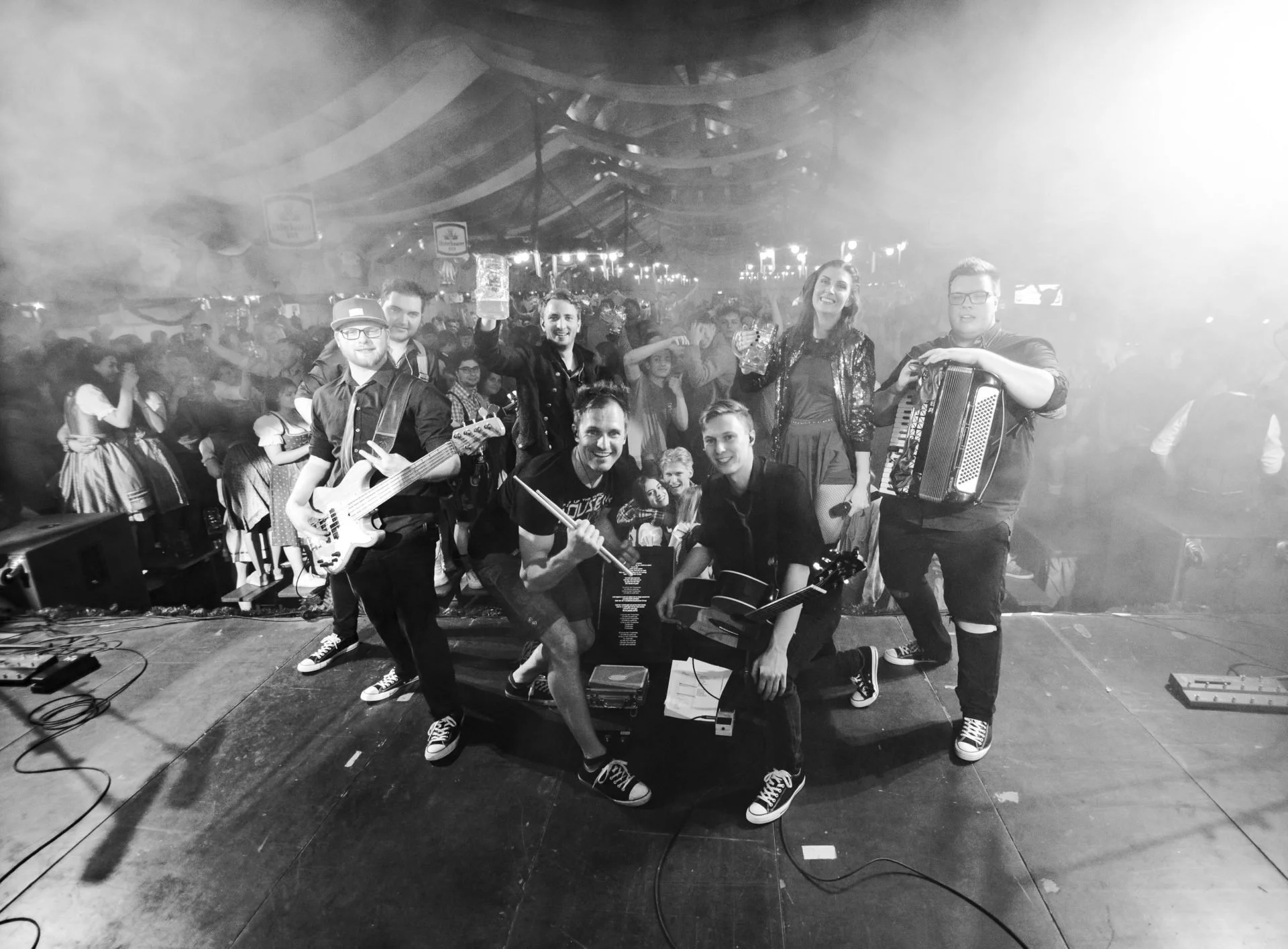 Eine Band mit sieben Mitgliedern auf einer Bühne bei einem Live-Konzert im Zelt. Sie spielen Instrumente und posieren für ein Foto, im Hintergrund ein Publikum, das festlich gekleidet ist.