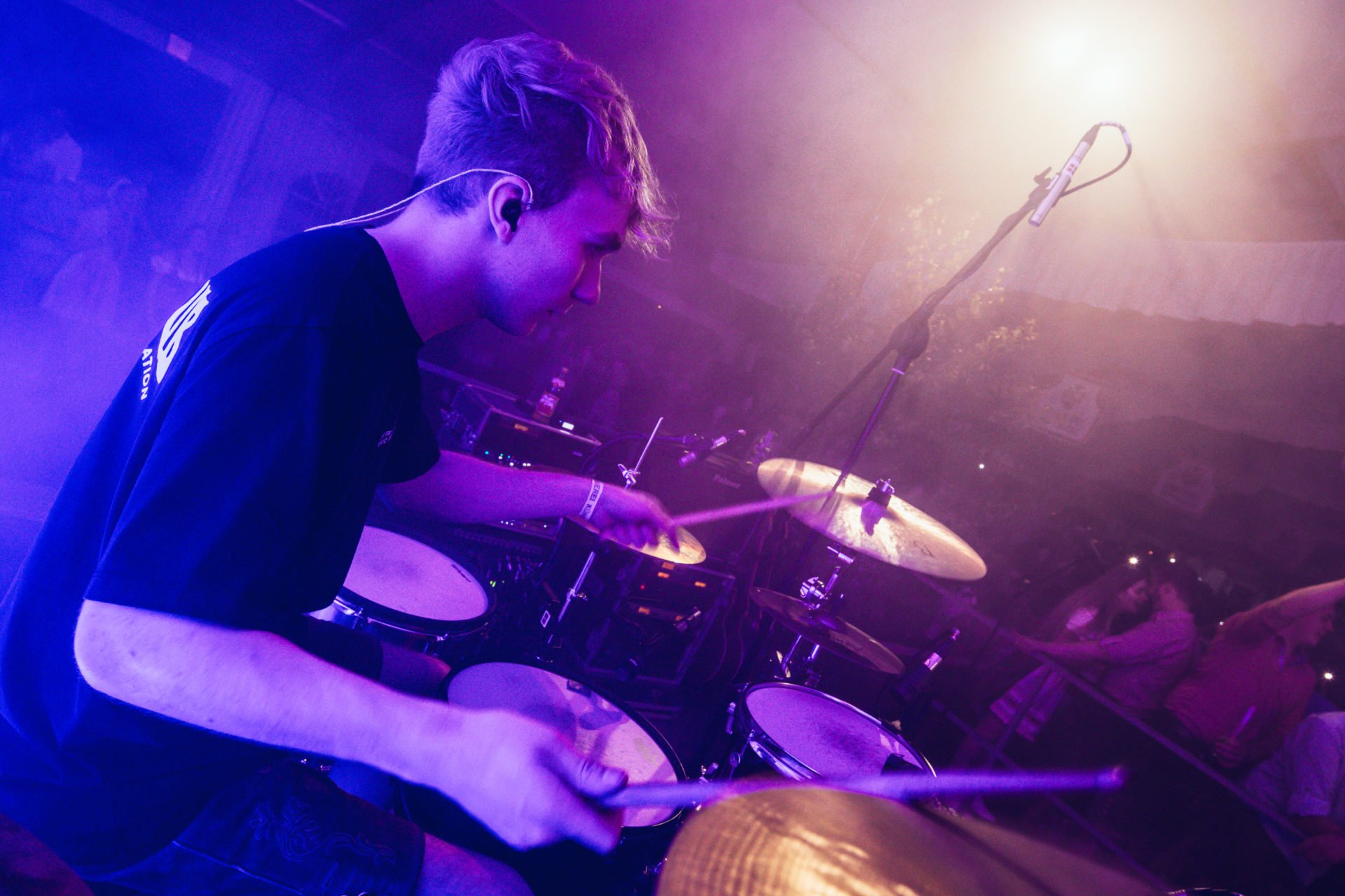 Ein junger Mann spielt Schlagzeug bei einer Live-Musikveranstaltung, mit buntem Bühnenlicht und einem Mikrofon im Hintergrund.