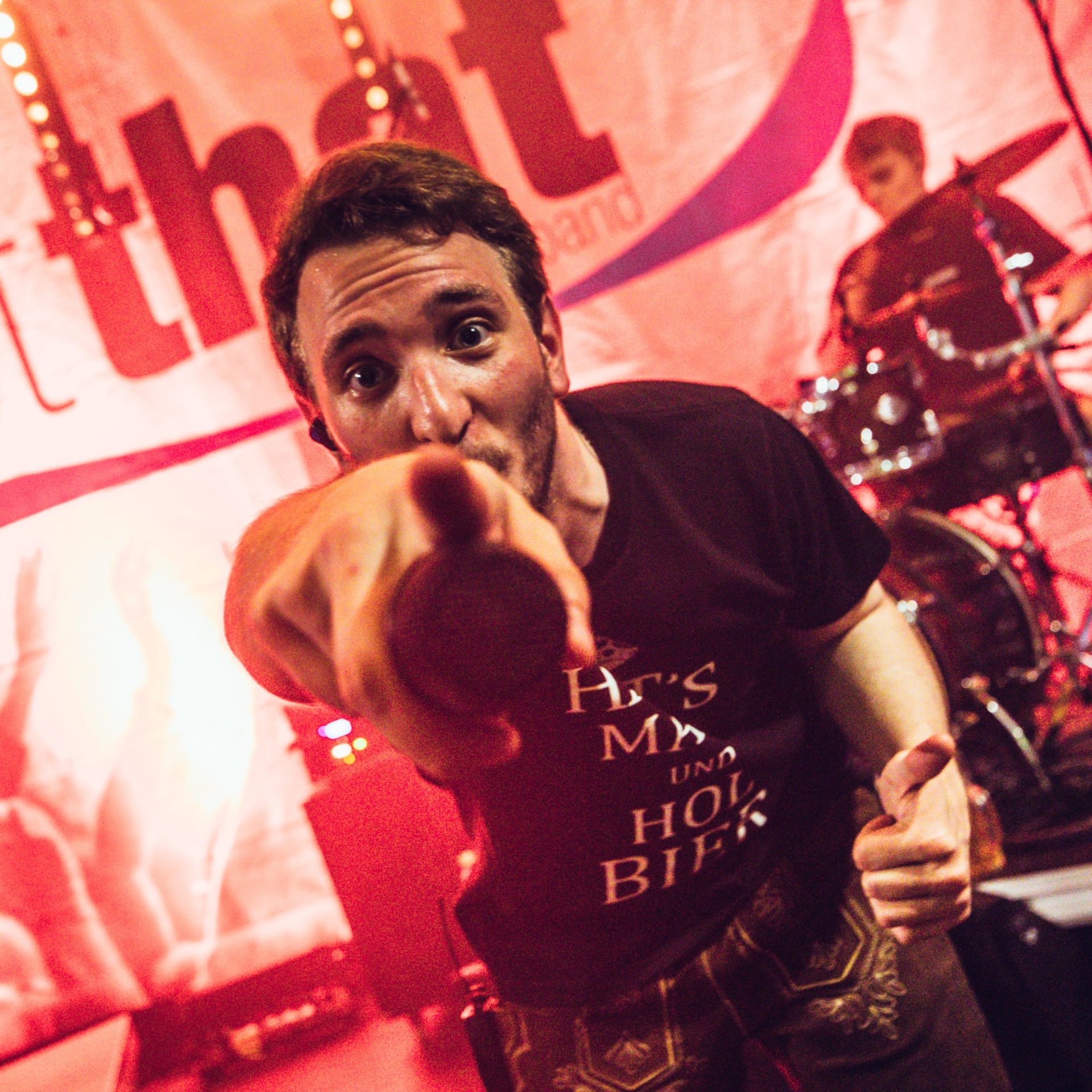 Ein Mann zeigt mit dem Finger in die Kamera, während er auf einer Bühne steht. Im Hintergrund ist ein Schlagzeug und eine Person, die Schlagzeug spielt, zu sehen.