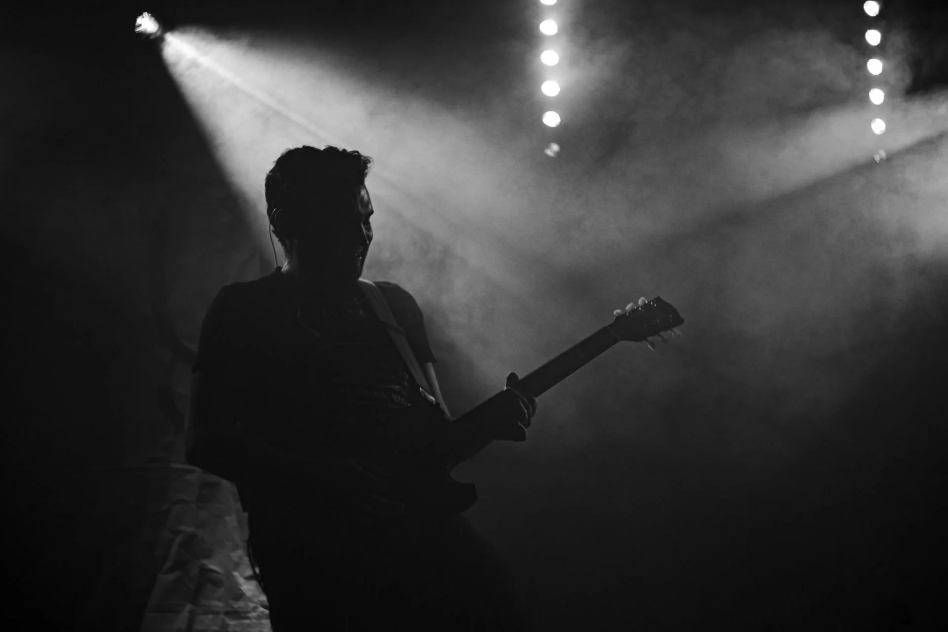 Eine Silhouette eines Gitarrenspielers auf einer Bühne mit Lichtstrahlen im Hintergrund, schwarz-weiß aufgenommen.