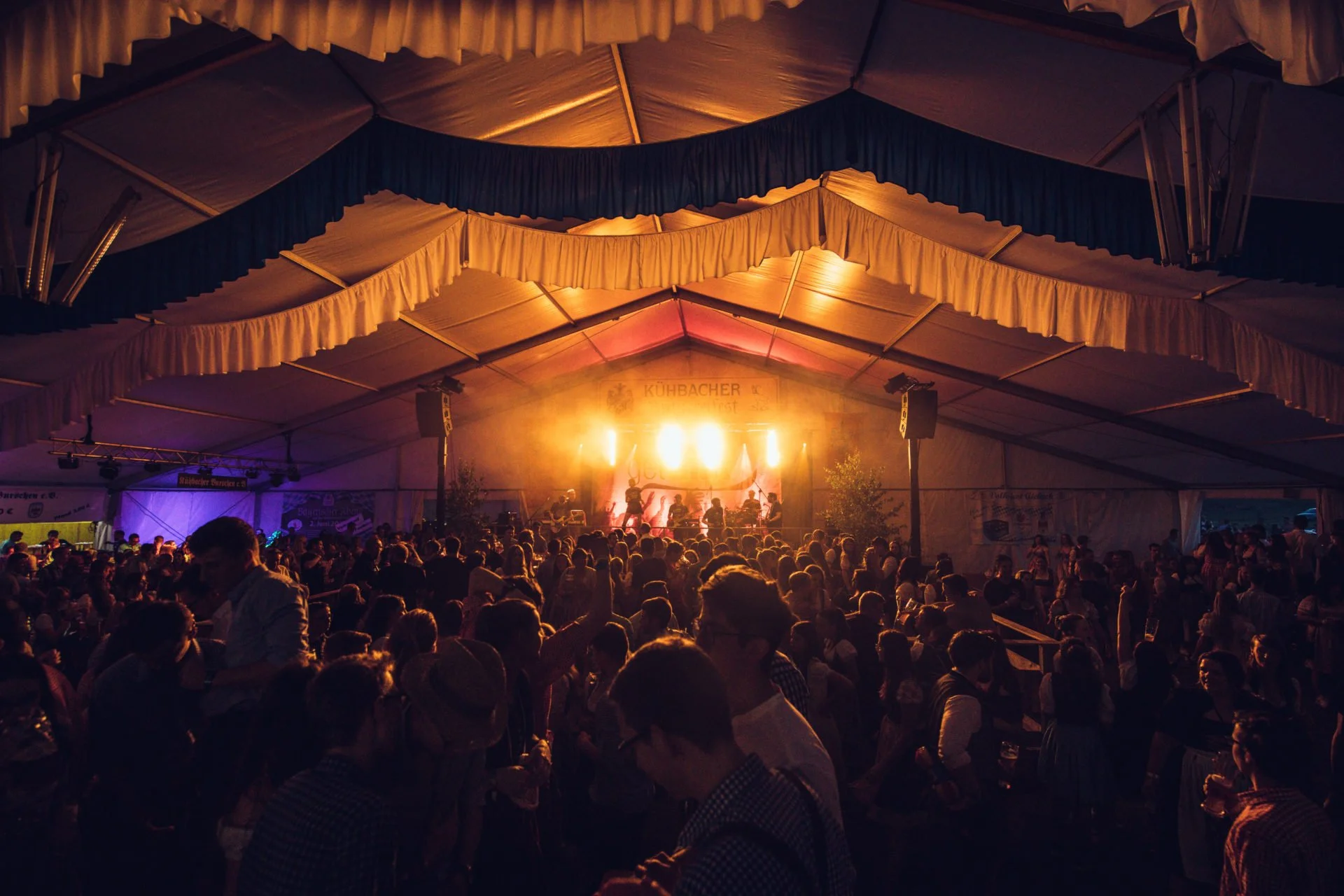 Menschen tanzen und feiern in einer großen Zeltveranstaltung mit Bühne, buntem Licht und Musik auf einem Volksfest.