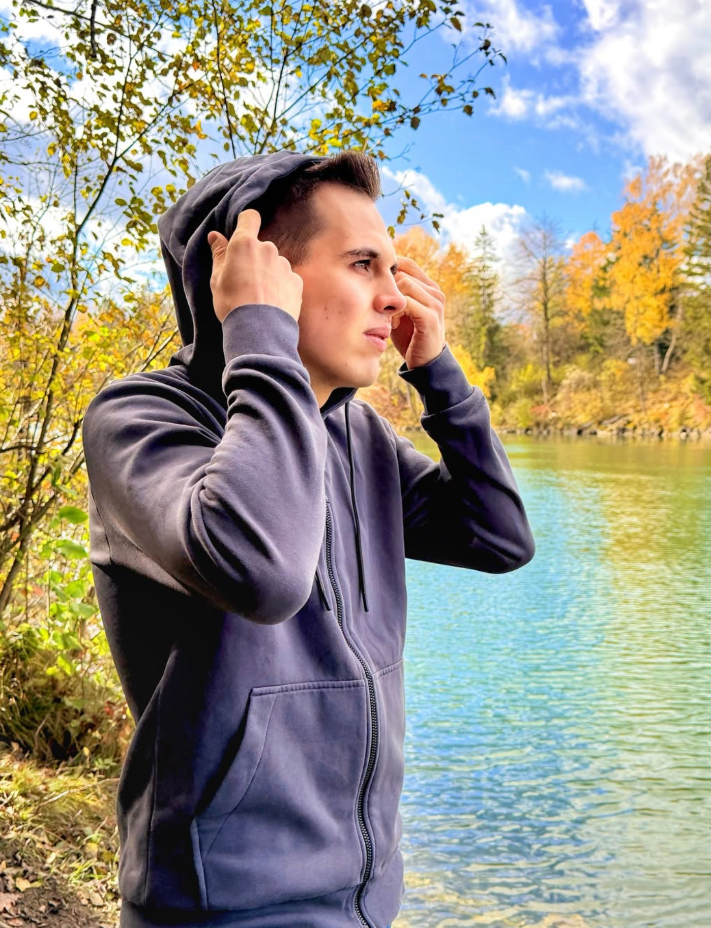 Junge Mann in grauem Kapuzenpullover steht am Ufer eines Sees, umgeben von Bäumen mit Herbstlaub, während er die Ohren mit den Händen bedeckt und in die Ferne blickt.