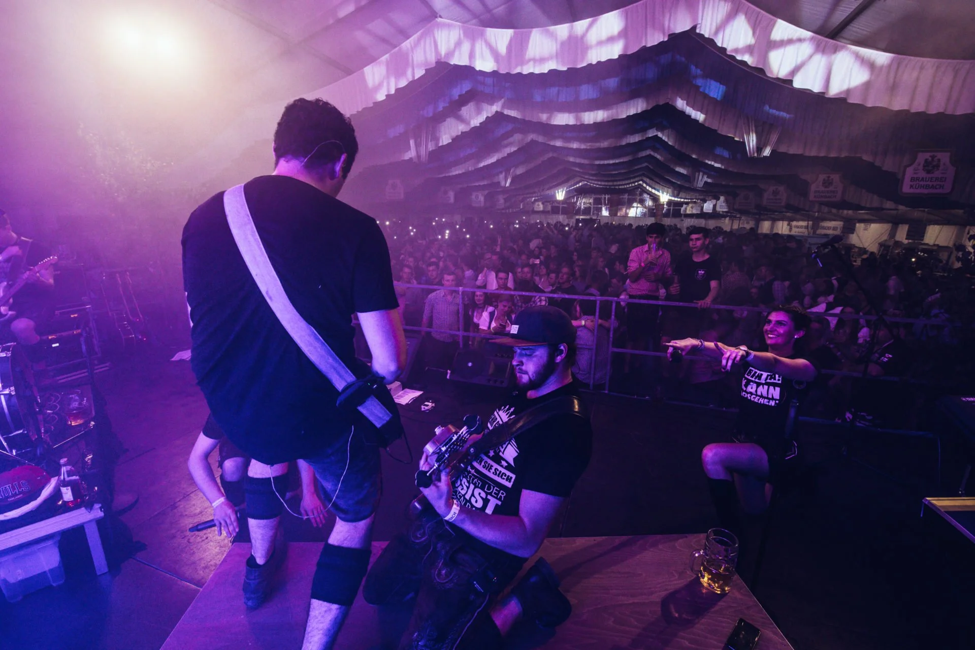 Junge Musiker auf Bühne während eines Konzerts in einer großen Festhalle, Publikum im Hintergrund, die Stimmung lebhaft und fröhlich, mit bunter Beleuchtung.