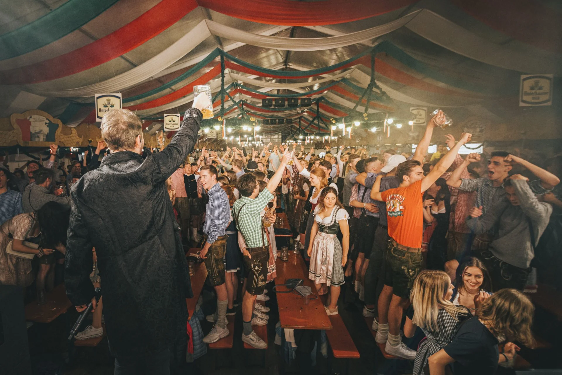 Viele Menschen in traditioneller Tracht feiern in einem großen festlich dekorierten Zelt, einige heben Bierkrüge, andere tanzen und lachen, während im Hintergrund Lichter leuchten.