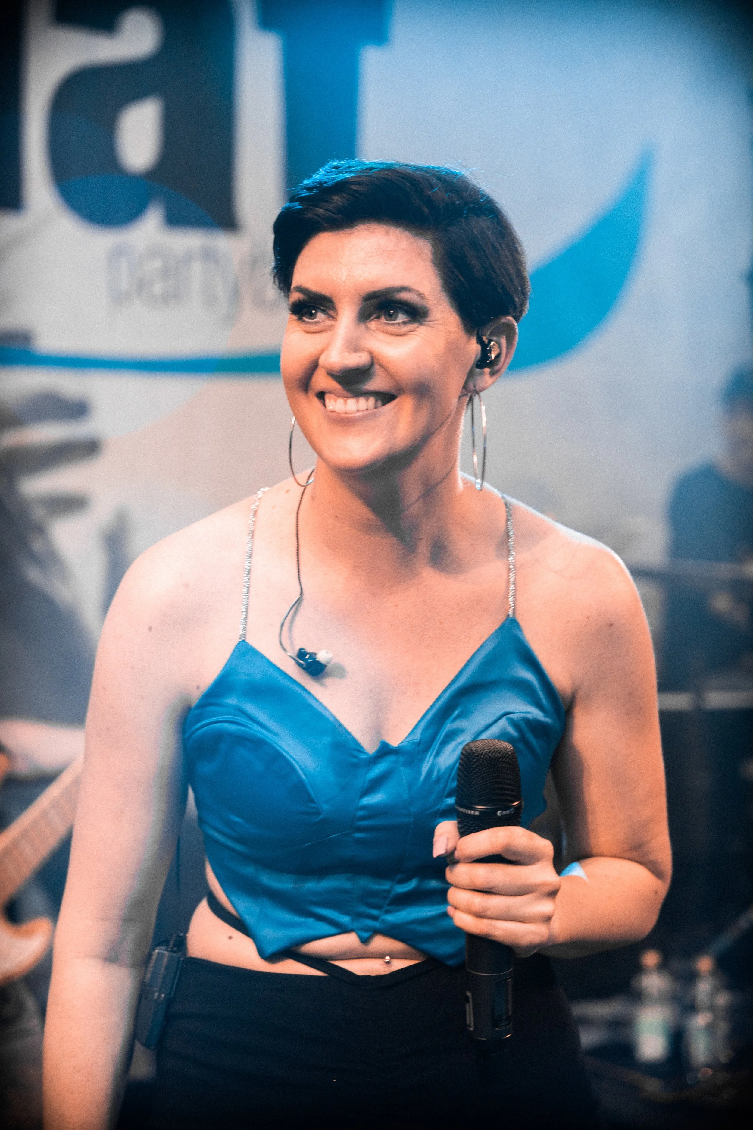 Eine lachende Frau mit Kurzhaarfrisur, die ein Mikrofon hält, trägt ein spitzenloses, blaues Top mit dünnen Trägern und großen Ohrringen. Sie steht auf einer Bühne bei einer Musikveranstaltung.